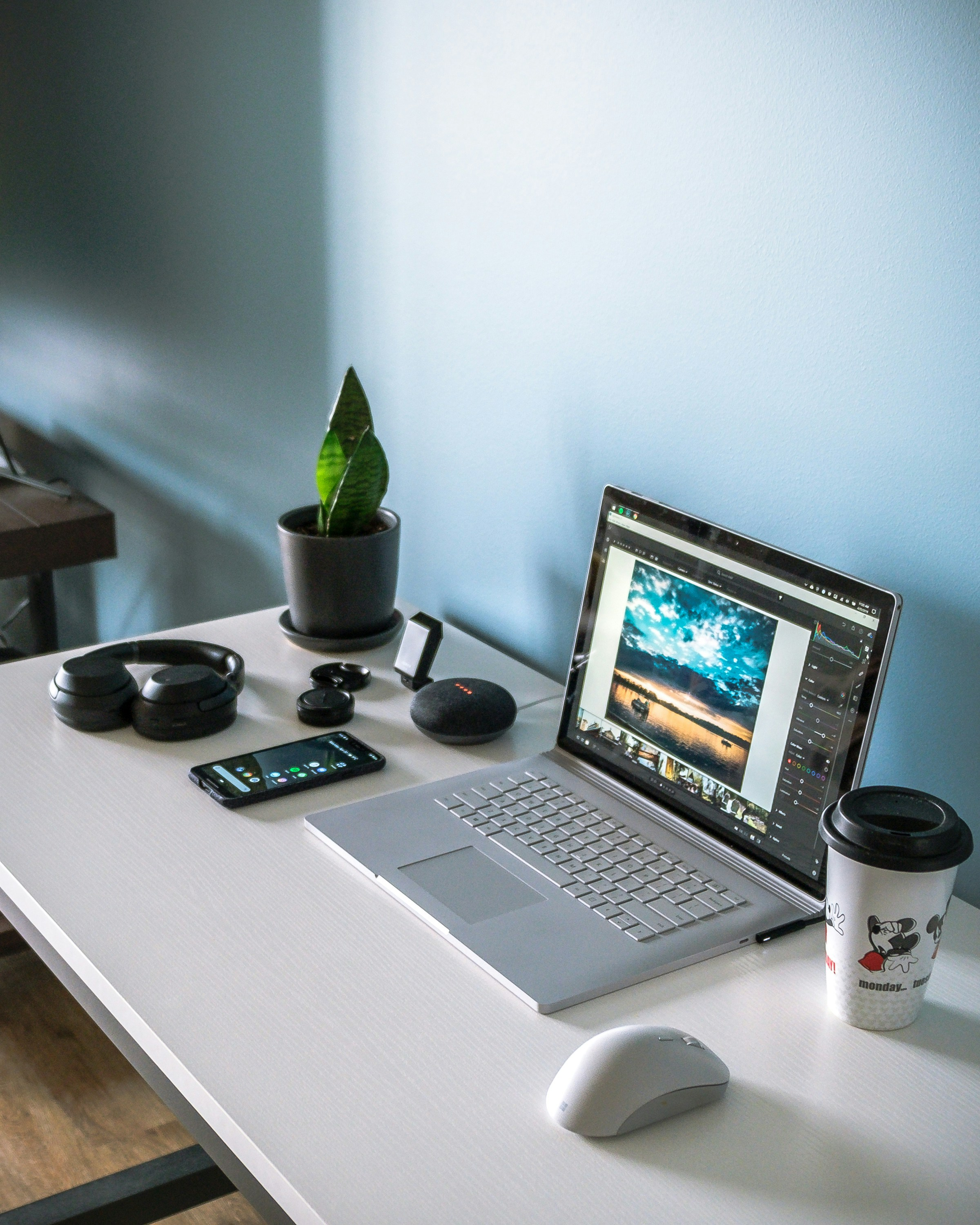 A white desk with a laptop displaying a landscape photo, a white computer mouse, a disposable coffee cup with Mickey Mouse design, a smartphone, a pair of wireless headphones, a digital voice recorder, a small potted plant, and a black smart speaker.
