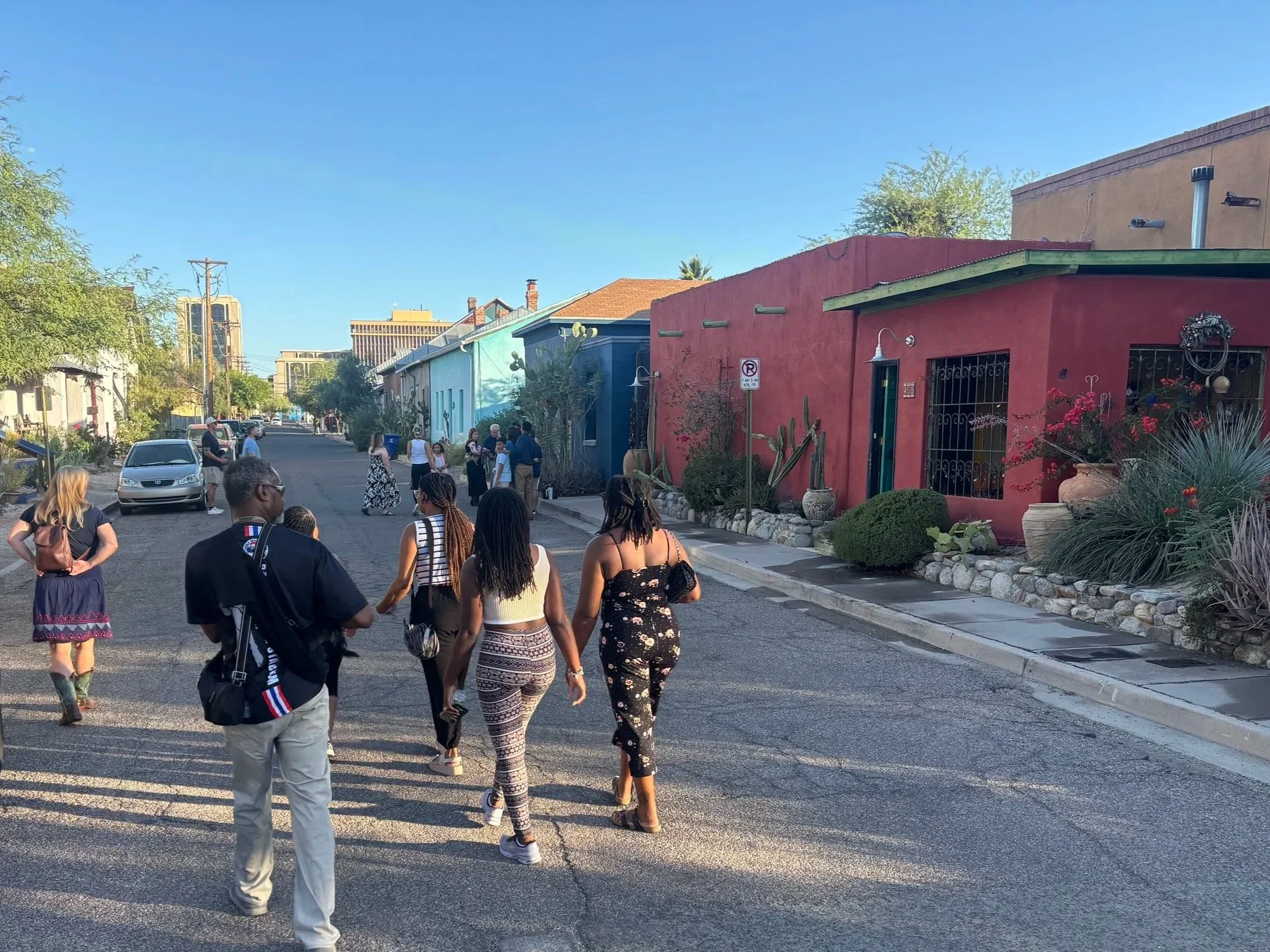 A group of people walking on a sidewalk in a colorful downtown Tucson neighborhood with painted houses, desert plants, and a clear blue sky.