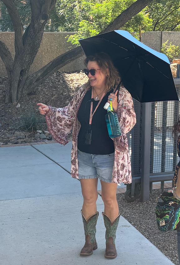 A woman with sunglasses holding a blue umbrella and a spray bottle, smiling outdoors on a sunny day, wearing a floral kimono, black top, denim shorts, and cowboy boots.