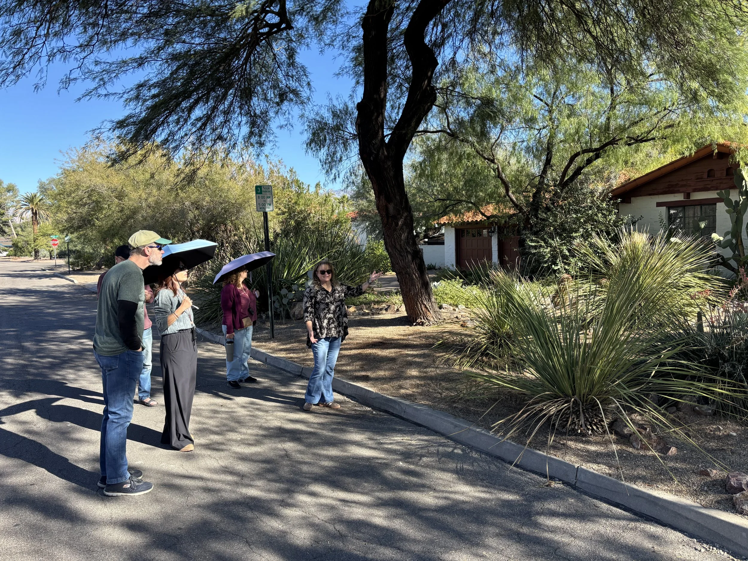 A group of five people standing on a sidewalk beside a landscaped yard with desert plants and trees, some holding umbrellas, on a sunny day.