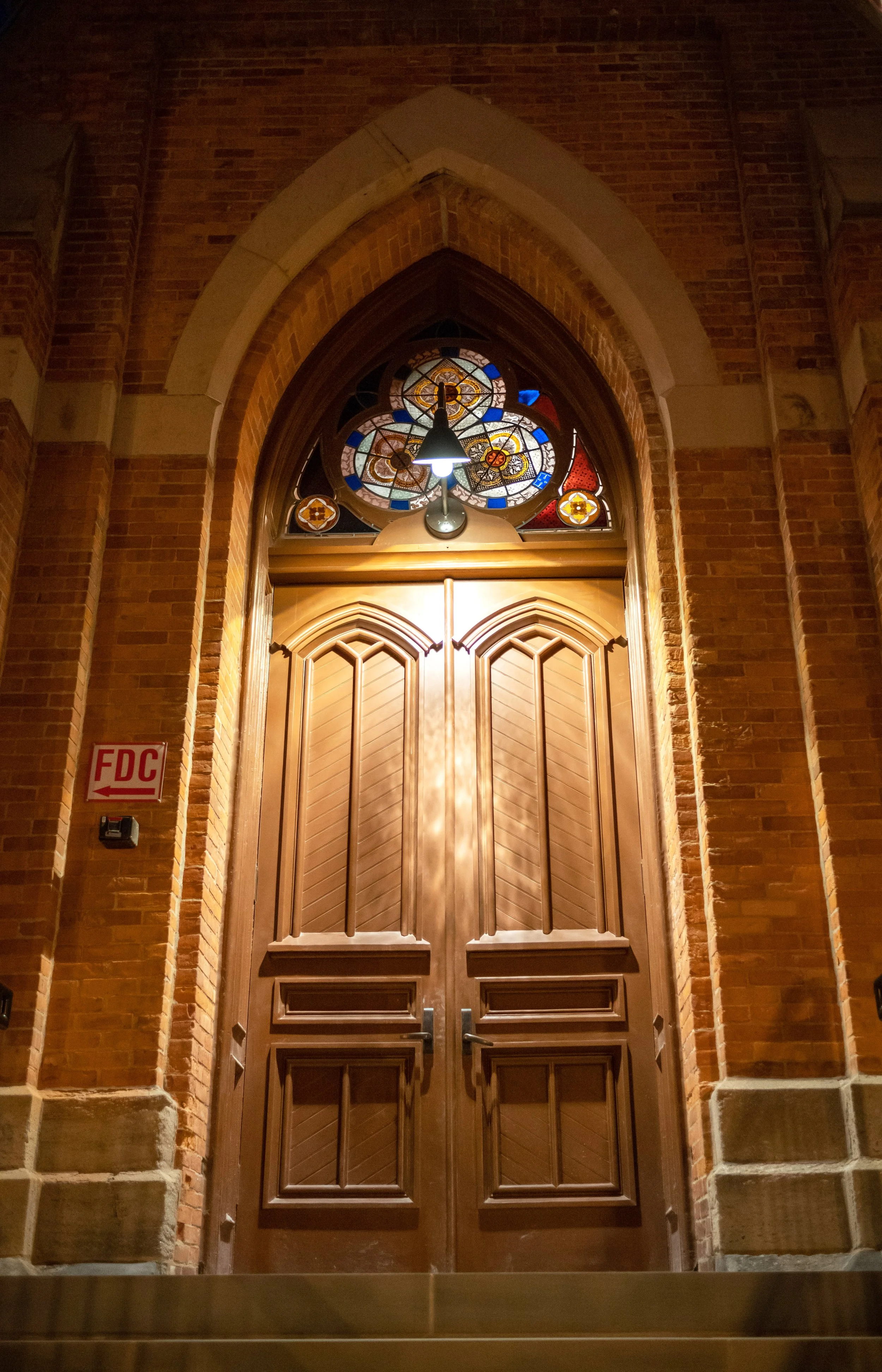 Large light warm brown wooden doors with pointed arch and trim detailing and a stained glass topper.