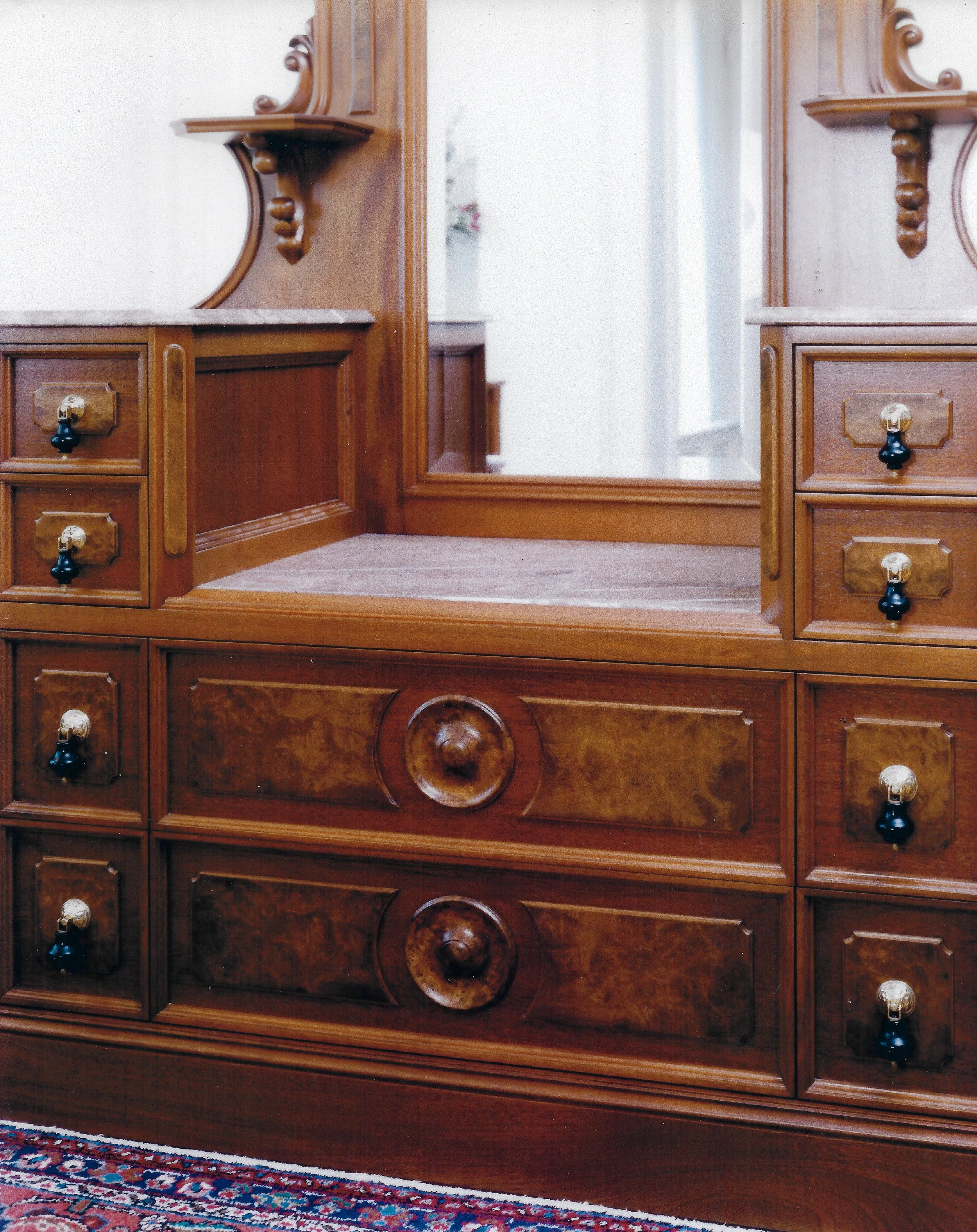 Close up on custom built, ornate, warm brown, mahogany, wooden, Victorian Era style dresser with dangling black knob pulls, and  a tall mirror built into it flanked by two small ornate display shelves.
