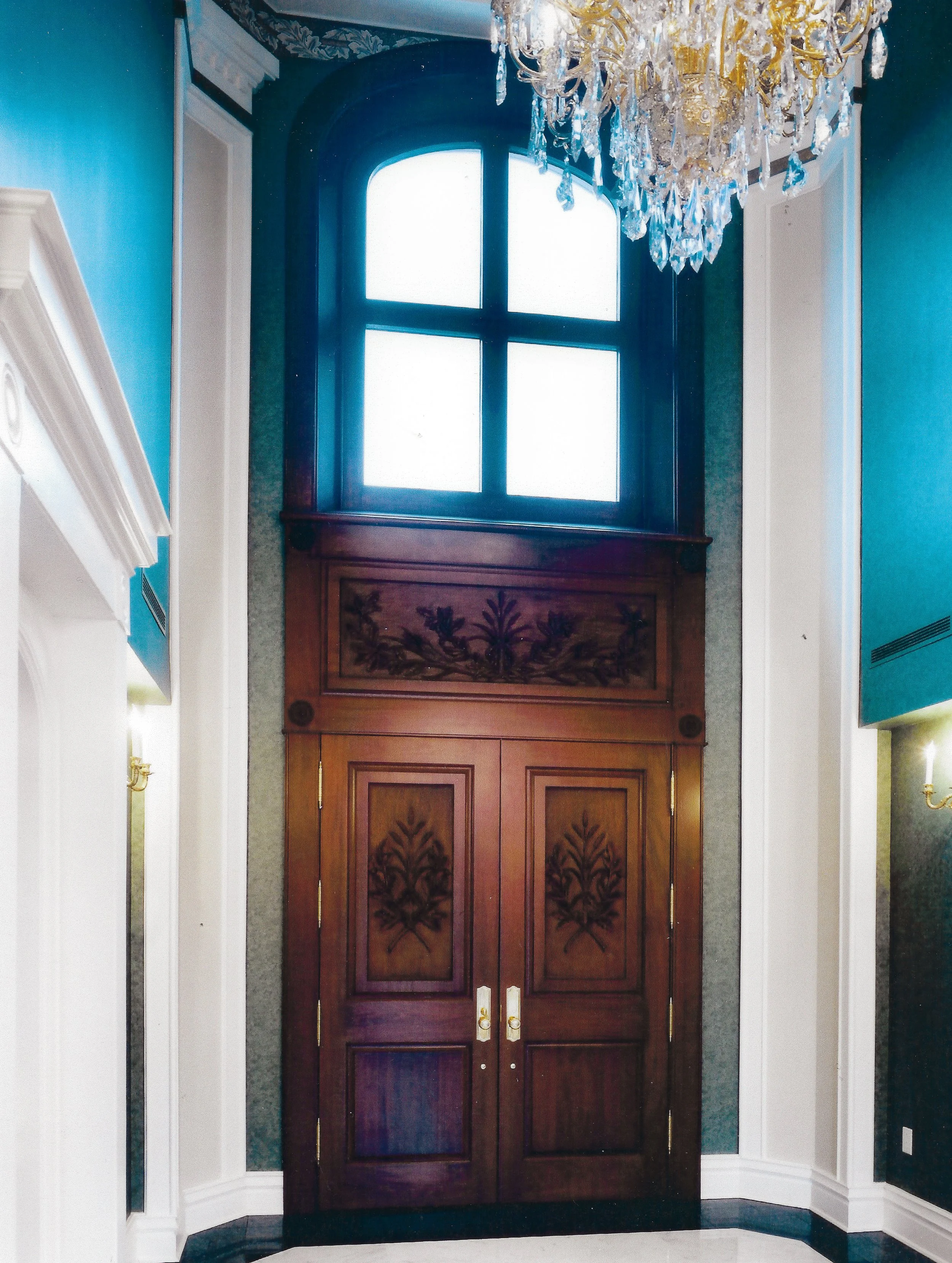 Tall dark brown mahogany wooden doors with a. large arched window above them and ornate carvings and trim details.