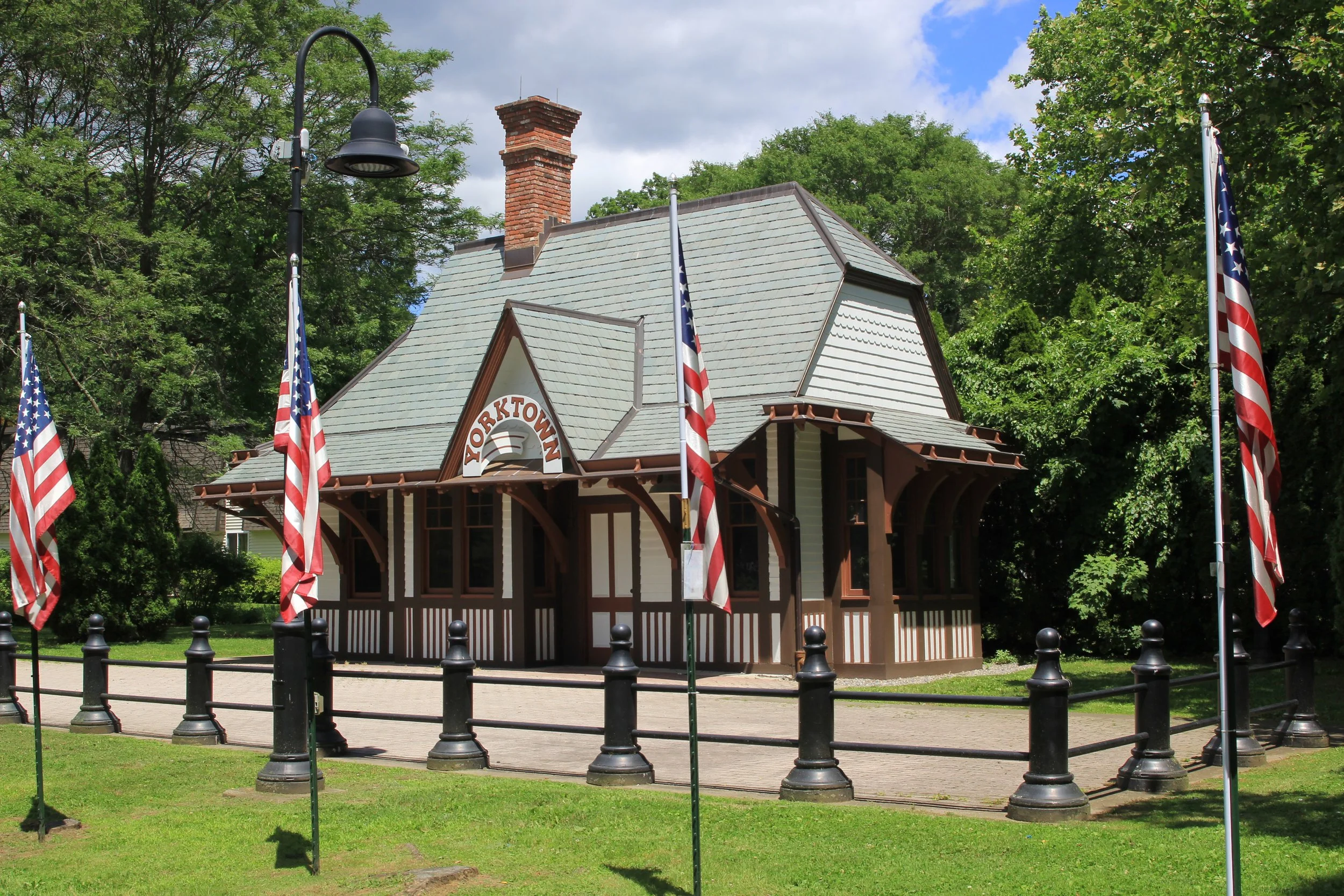 Small building with an combination gamble style light green roof, cream colored siding, dark brown wooden half-arch supports and detail elements, and a curved sign that reads Yorktown.