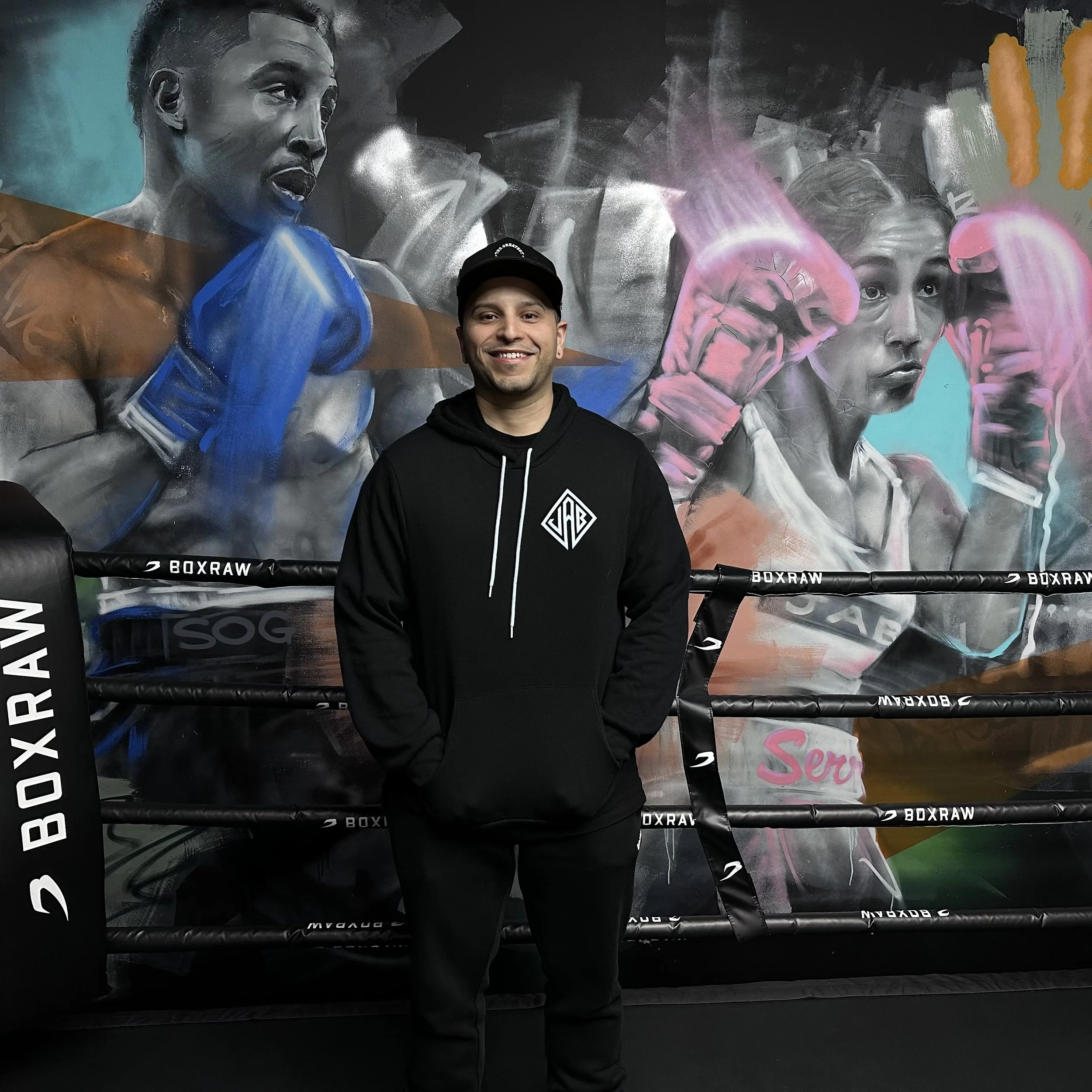 A young man stands in a narrow alleyway between beige and red brick buildings, wearing a gray blazer, black shirt, black pants, and white sneakers, with hands in his pockets, looking at the camera.