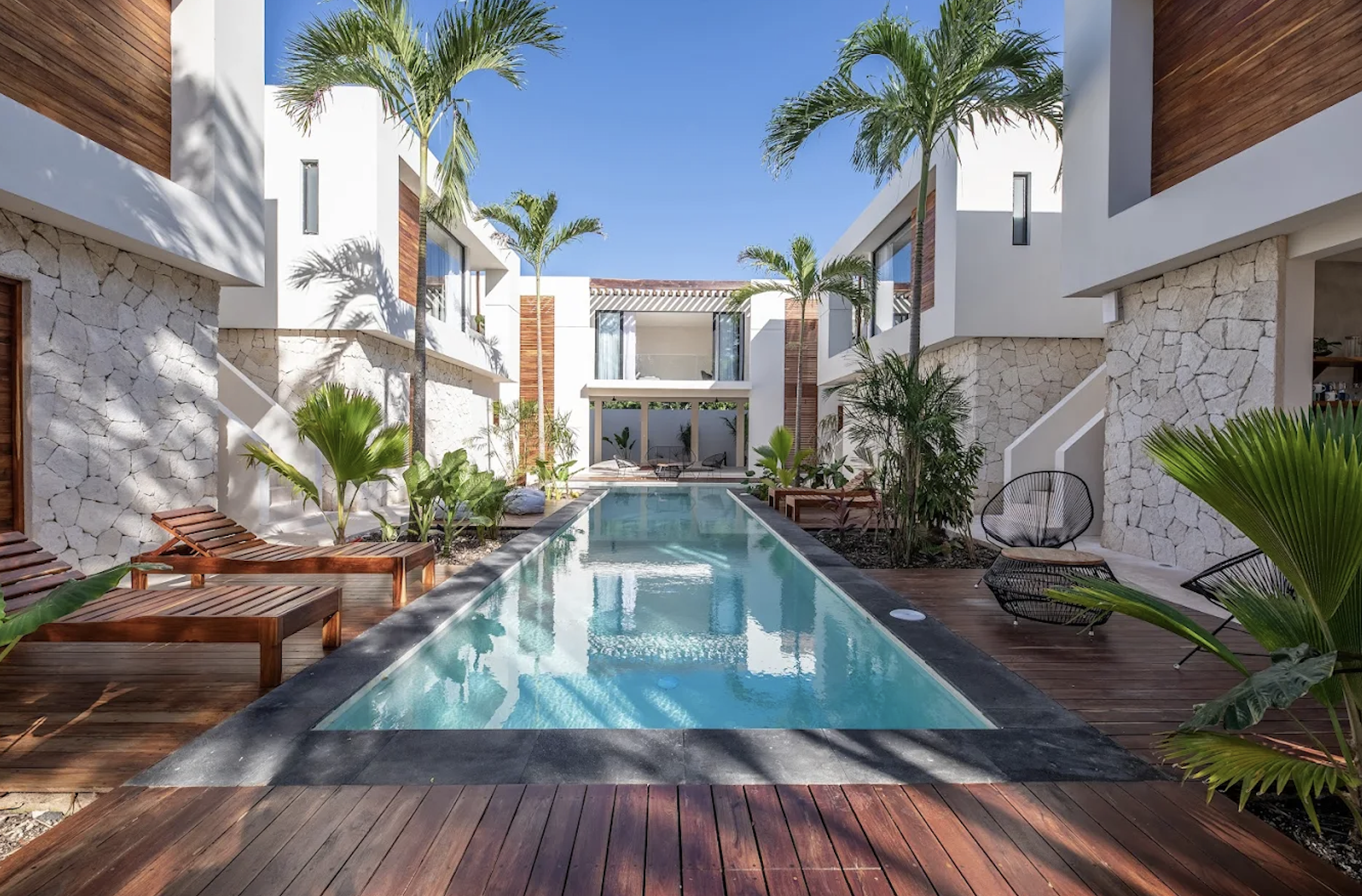 A modern outdoor pool area with wooden deck, lounge chairs, tropical plants, and surrounding white buildings with wood accents and large windows, under a clear blue sky.