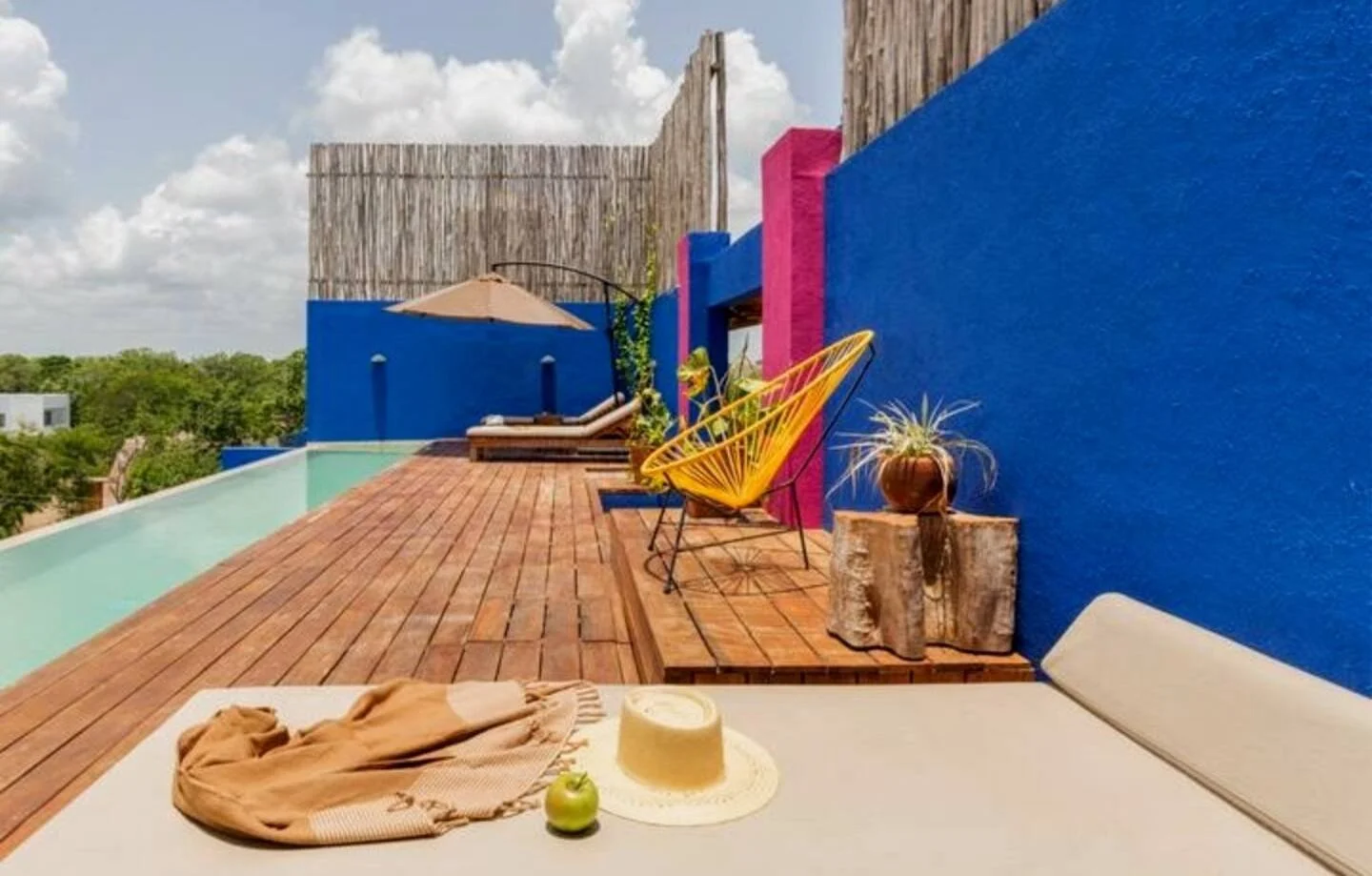 Colorful rooftop patio with wooden deck, colorful walls, sun hat, apple, and lounge furniture under a cloudy sky.