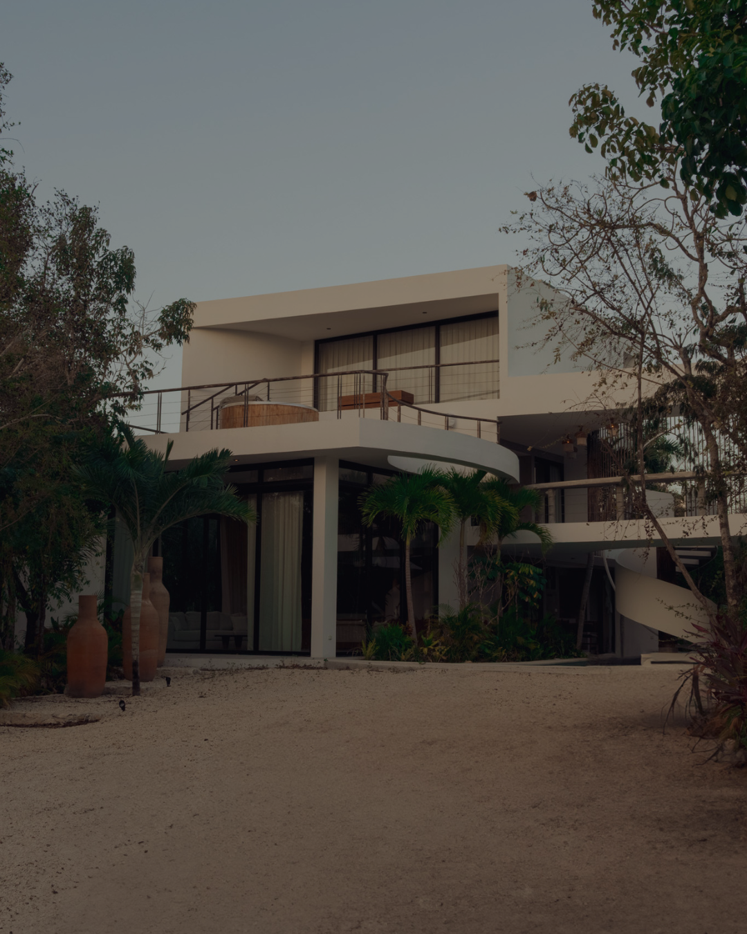 Modern white multi-level house with curved balcony, surrounded by trees and desert landscape, under a clear sky.