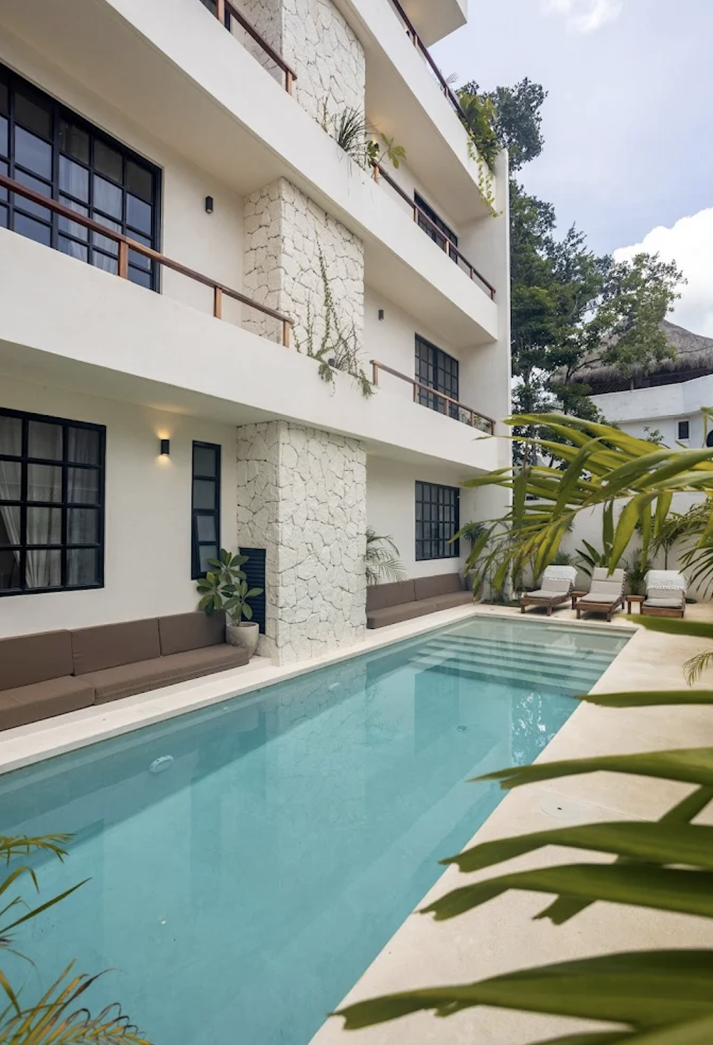 Modern hotel courtyard with a rectangular swimming pool, surrounded by lounge chairs, cushioned seating, and tropical plants, with a white multi-story building in the background.