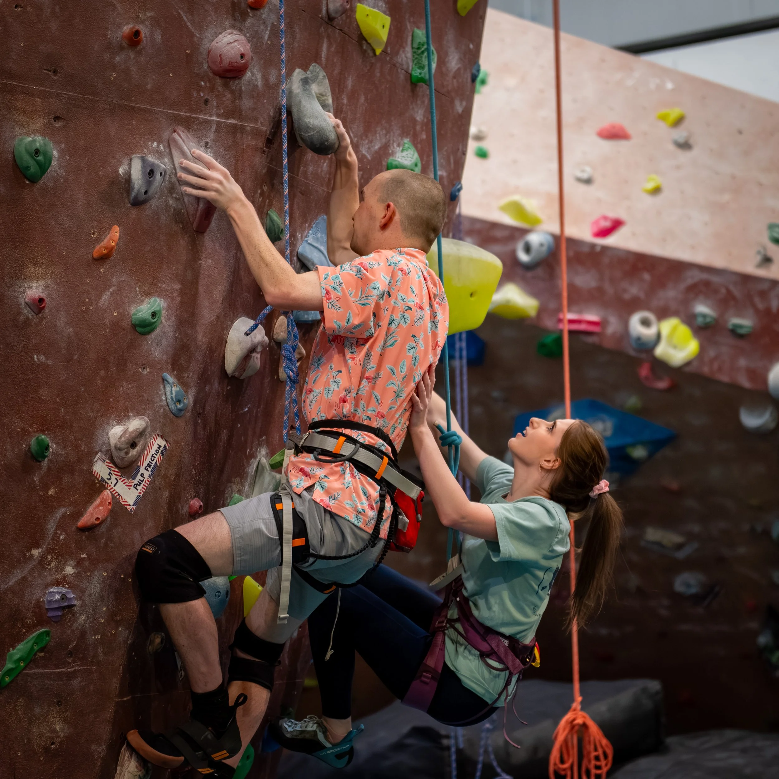 Sophia Barondeau side-climbs with Chuck Winstead, who is blind, at Rocksport Climbing Gym. Chuck is wearing an orange Hawaiian shirt and knee pads. Sophia helping and climbing next to him sports a tight ponytail and a look of concentration.