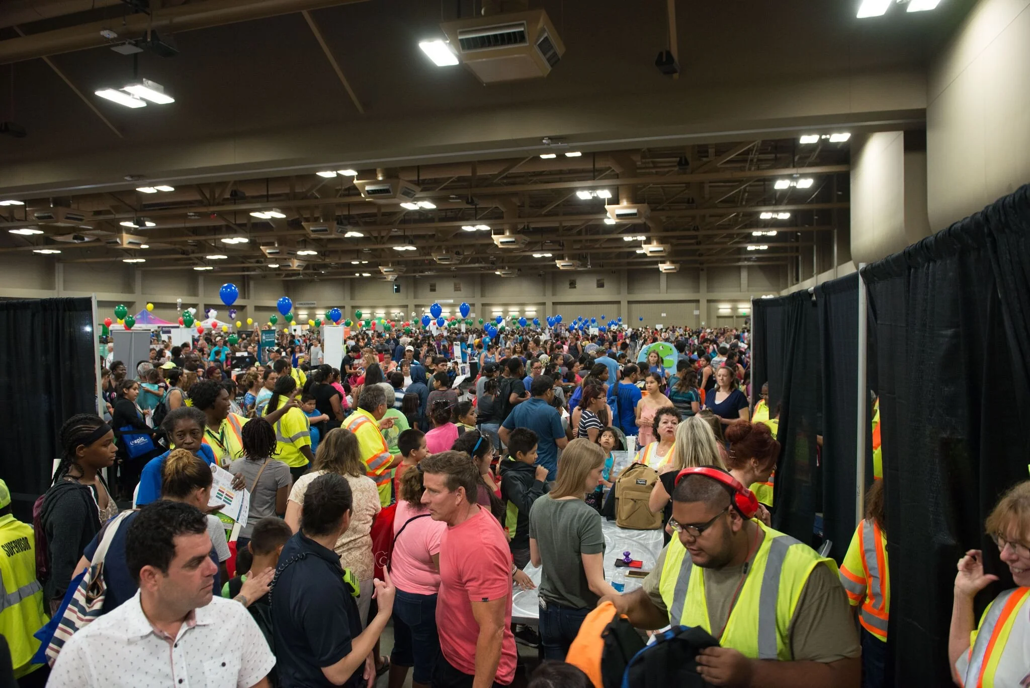 Crowd attending a large indoor event with booths, colorful balloons, and many people wearing bright yellow safety vests.