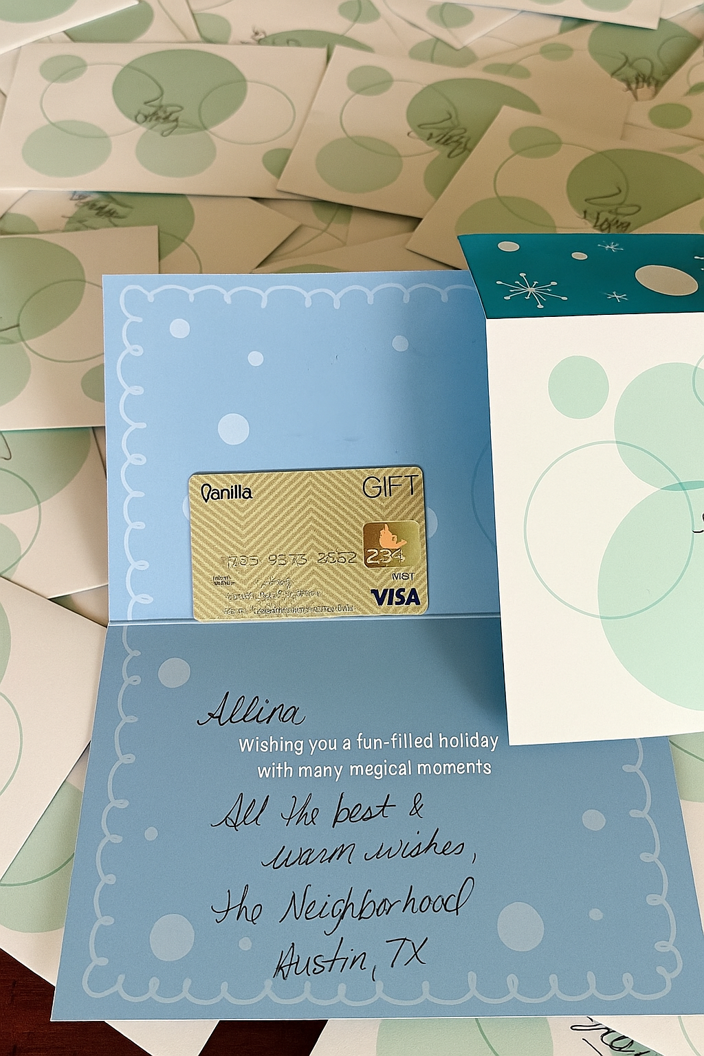 A light blue holiday card with white polka dots and a handwritten message inside. The message reads, "Allina, Wishing you a fun-filled holiday with many magical moments. All the best & warm wishes, The Neighborhood, Austin, TX." There is a gift card inside the card, which appears to be a Visa gift card, with some text and images on it. The background shows more holiday cards with a green circle pattern.