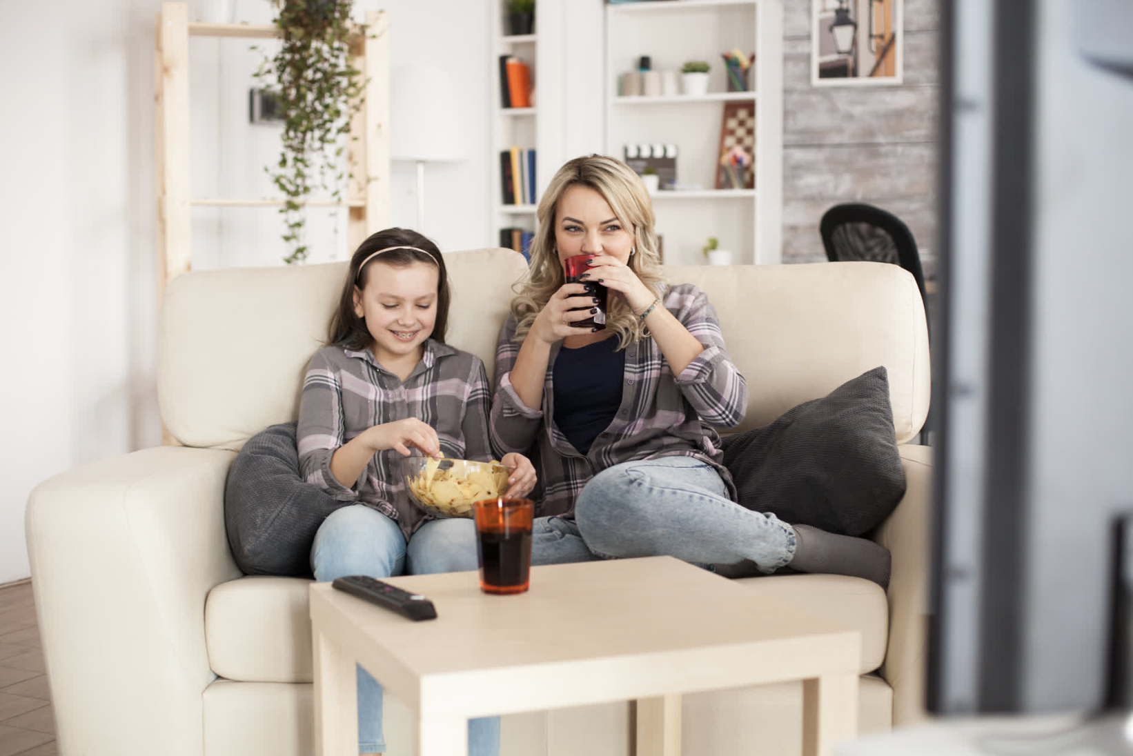 A woman and a girl sit on a beige couch watching TV, sharing snacks and drinks in a cozy living room with bookshelves and plants.