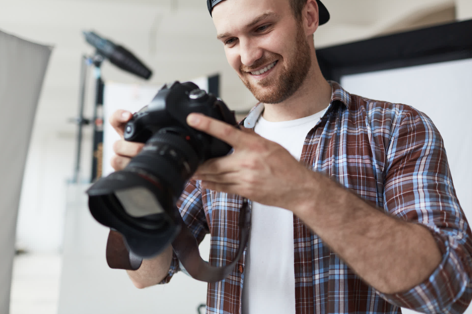 Young man in plaid shirt smiling and looking at camera in hands, indoors with photography equipment around.