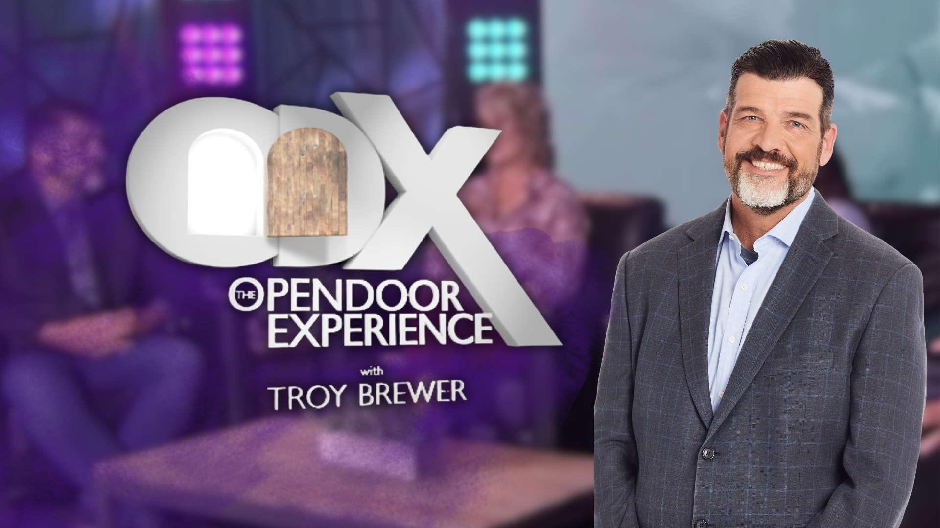 Troy Brewer smiling in a checkered blazer and light blue shirt stands in front of a blurred background featuring the logo of 'The Opendoor Experience with Troy Brewer,' with purple lighting and a woman sitting at a table.