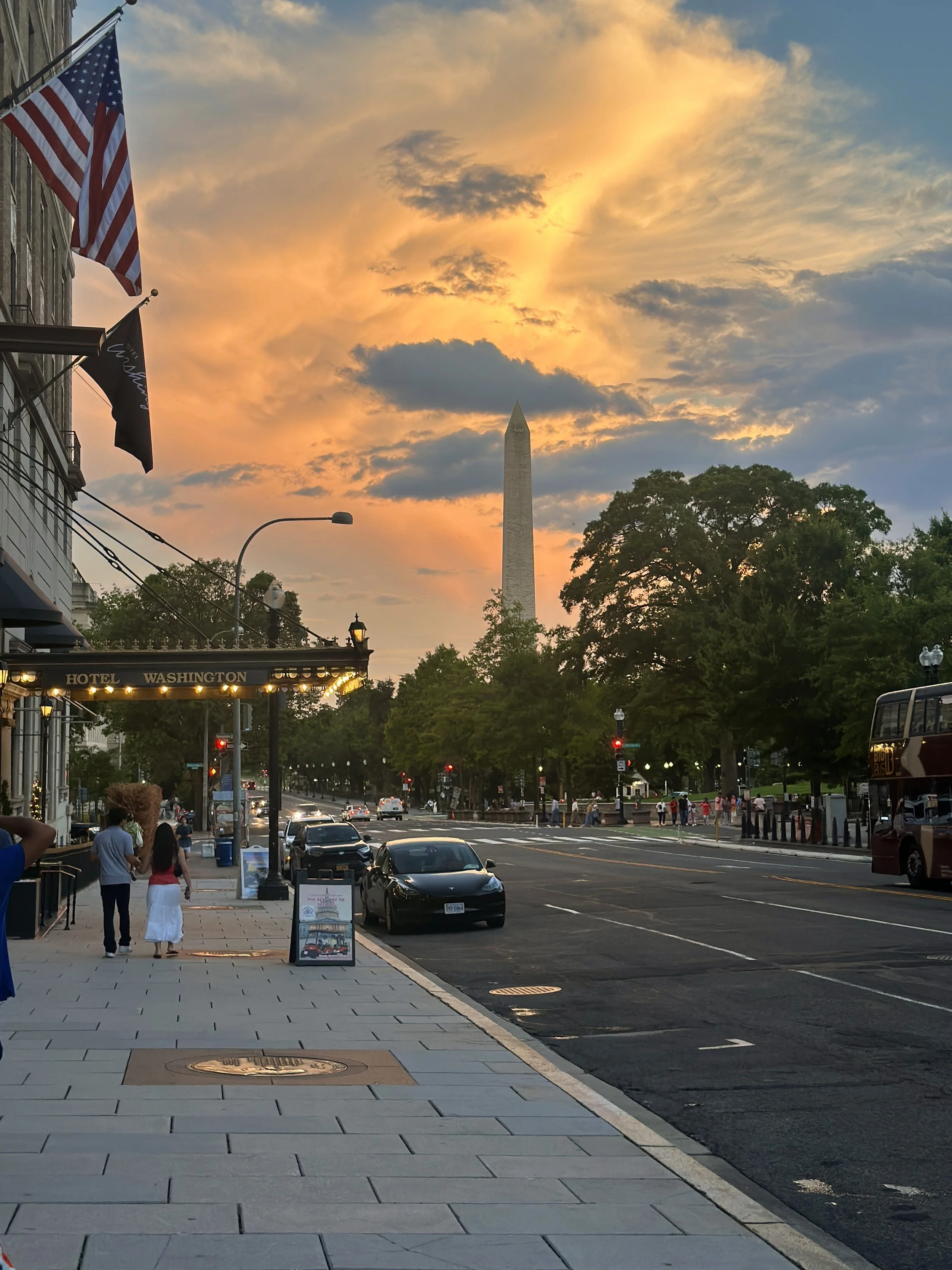 Sunset over a street in Washington D.C., featuring the Washington Monument, American flags, cars parked along the street, and people walking on the sidewalk.