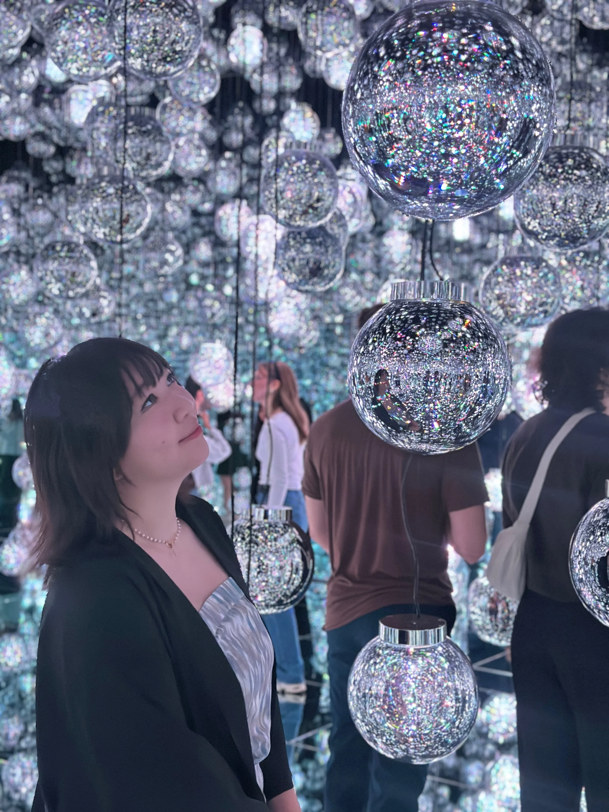 A woman is looking up at hanging reflective, glittering spheres in an art installation, with other visitors in the background.