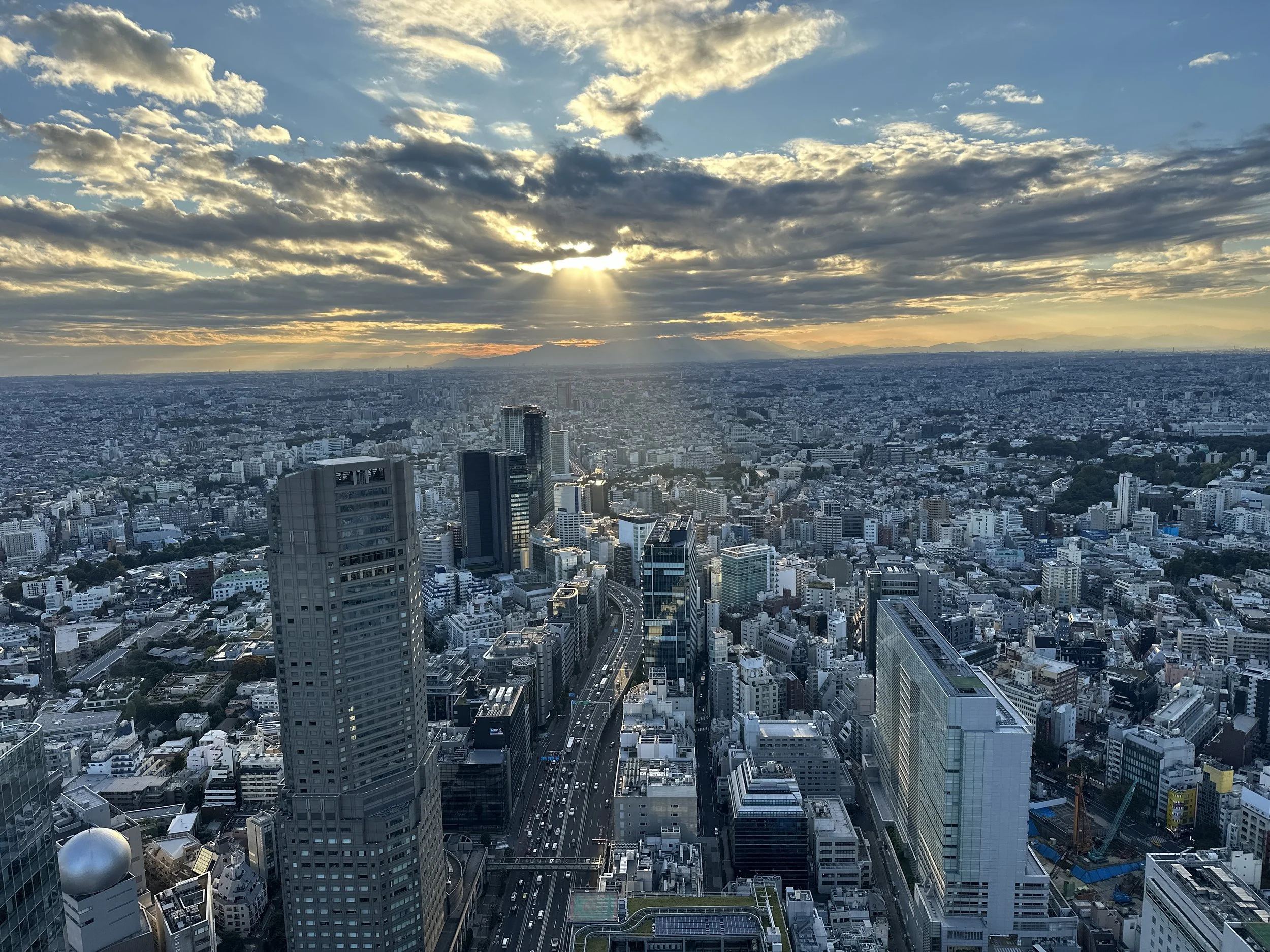A city skyline at sunset with skyscrapers and a highway, with sun rays breaking through clouds over distant mountains.