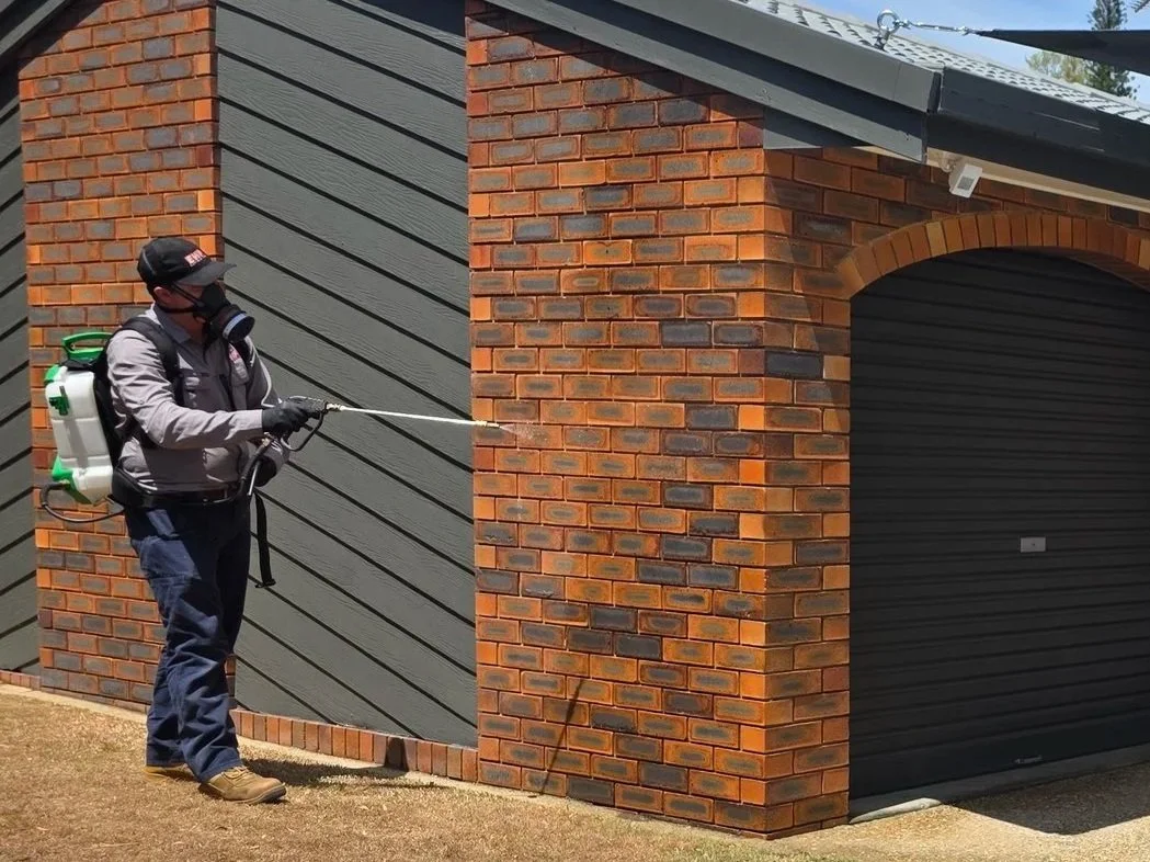 An East West Pest technician in protective gear using a backpack sprayer to disinfect the exterior brick wall of a building as part of Ipswich pest spray and inspection services..