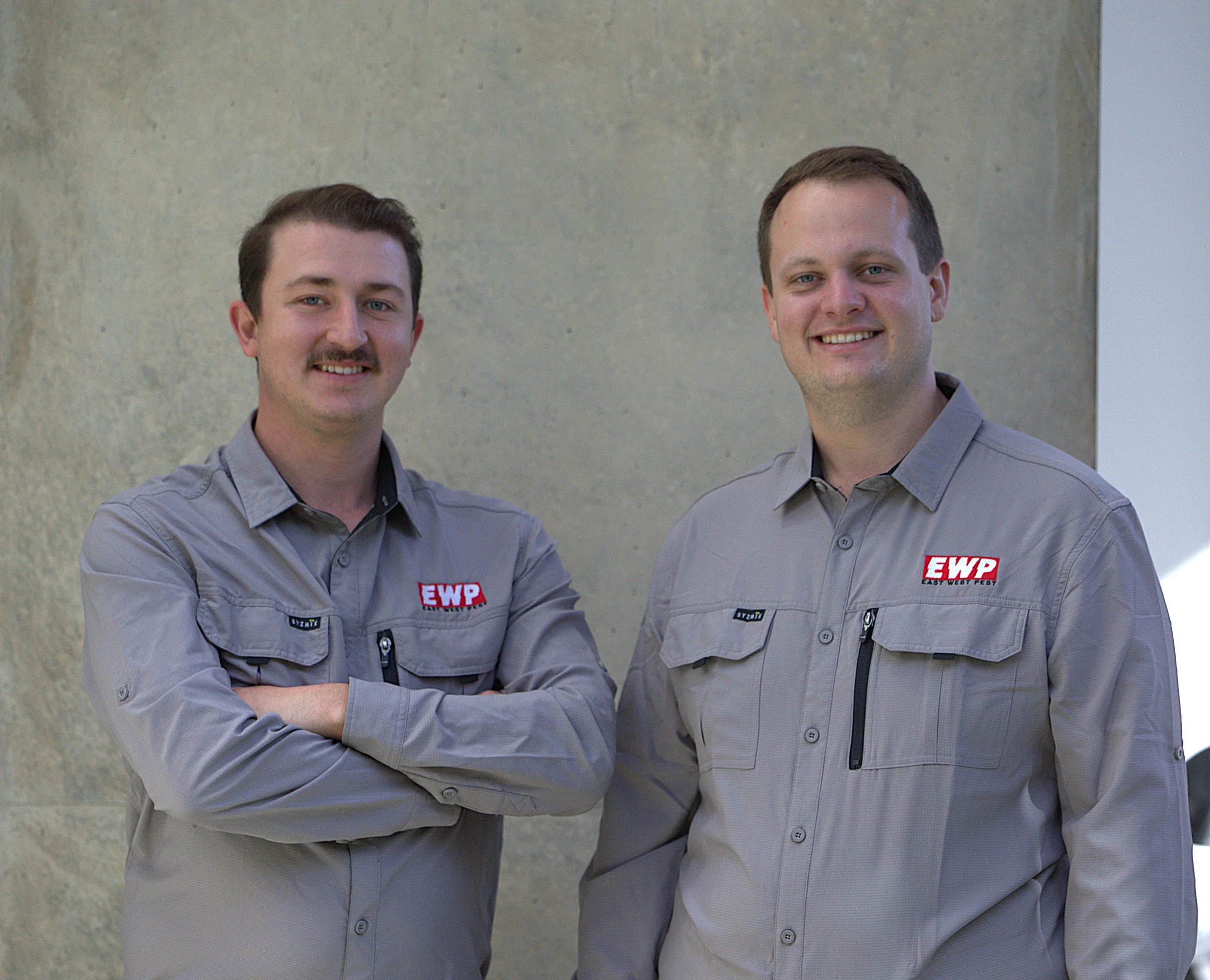 Ben and Andrew, professional Ipswich pest control technicians from East West Pest, smiling in grey EWP uniforms against a concrete wall.