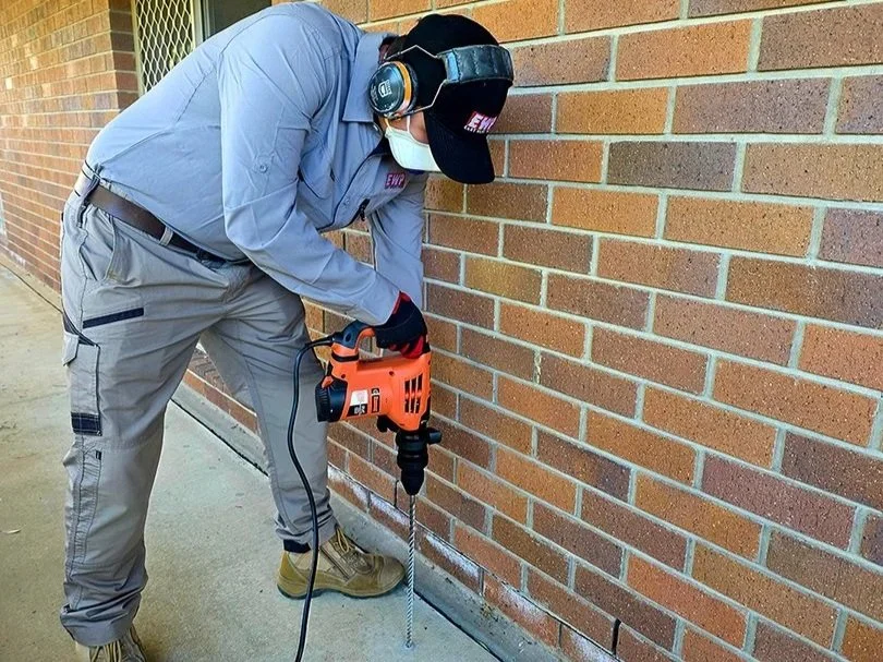 A technician drilling into a concrete slab around the external wall of a home so termite killing liquid spray can be injected and prevent termite damage to the house.