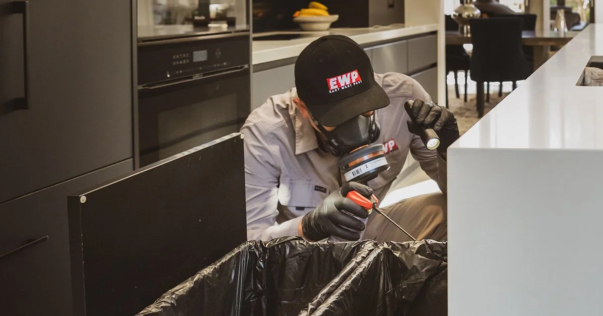 A man wearing a gray uniform, Black gloves, a gas mask, and a cap with a red and white logo, is performing a pest inspection, looking for cockroaches near a bin with a flashlight and pest spray can in a modern kitchen.