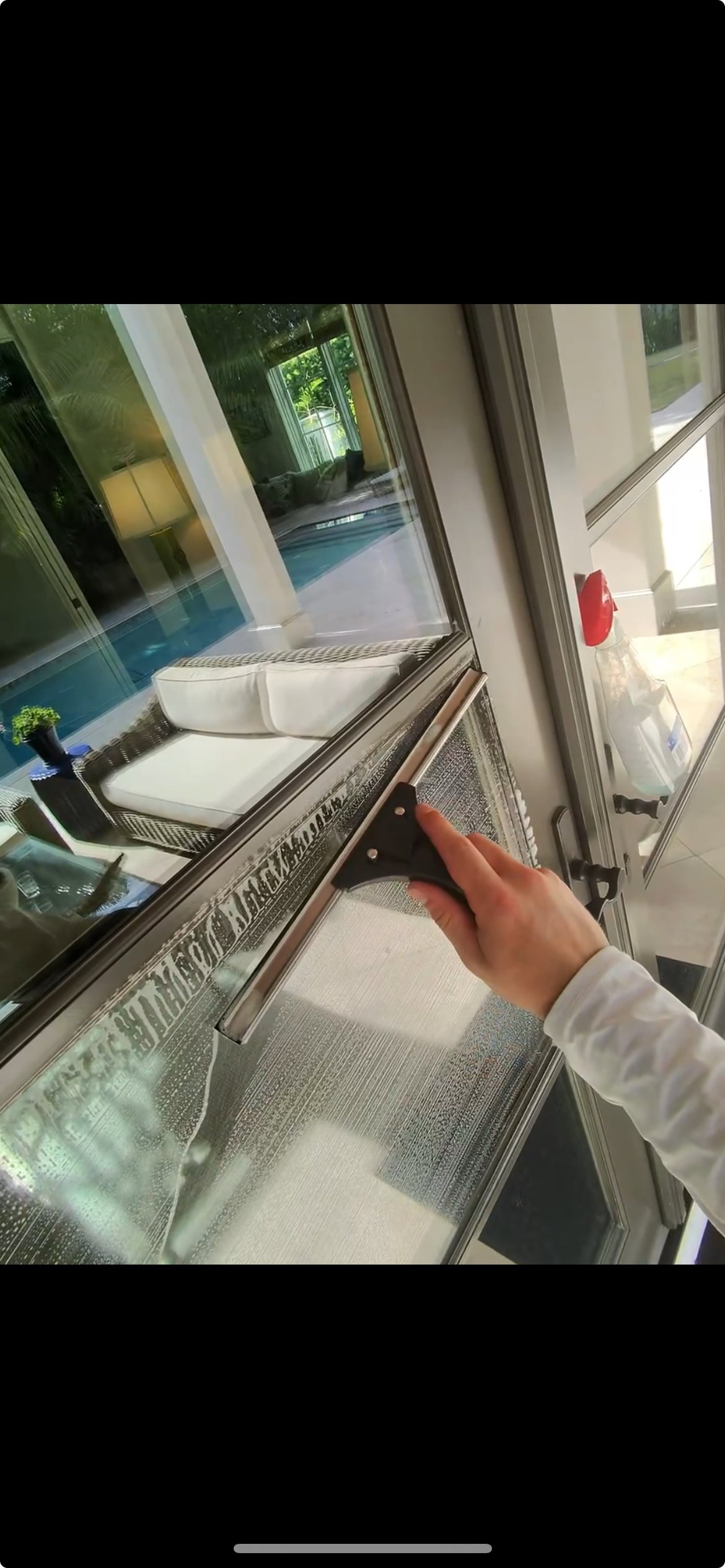 Person cleaning glass door with a squeegee, outdoor patio area with furniture and potted plant visible through the glass.