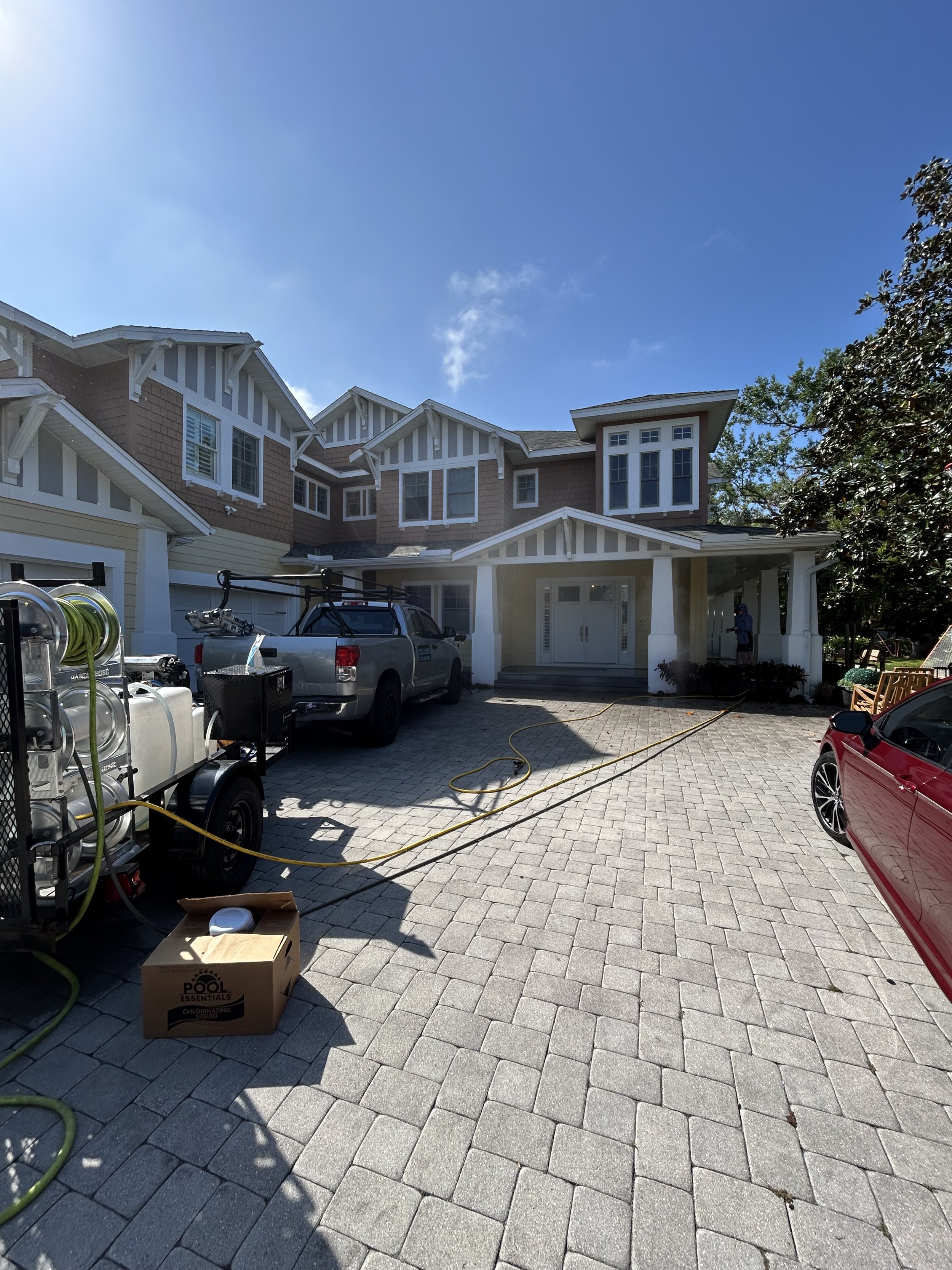 House with driveway, parked vehicles, and equipment for pool cleaning, with blue sky and some trees in the background.