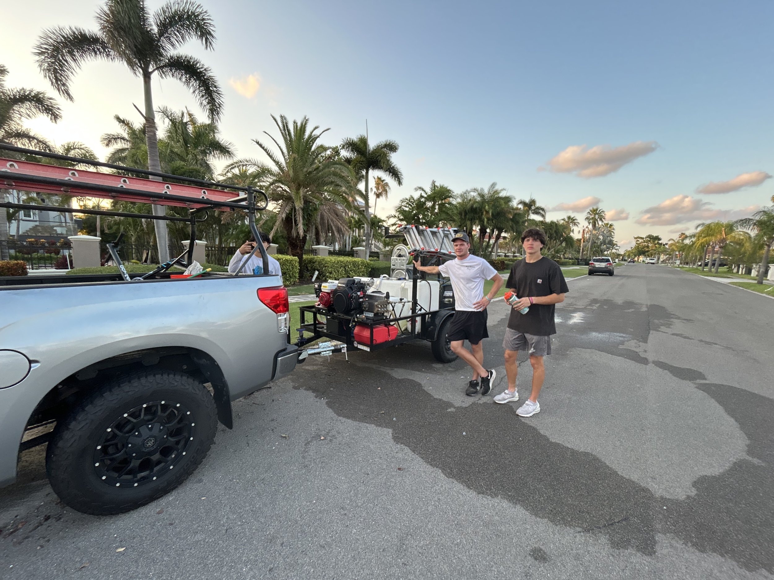 Two young men standing next to a trailer hitched to a gray pickup truck on a suburban street with palm trees, holding drinks, during sunset