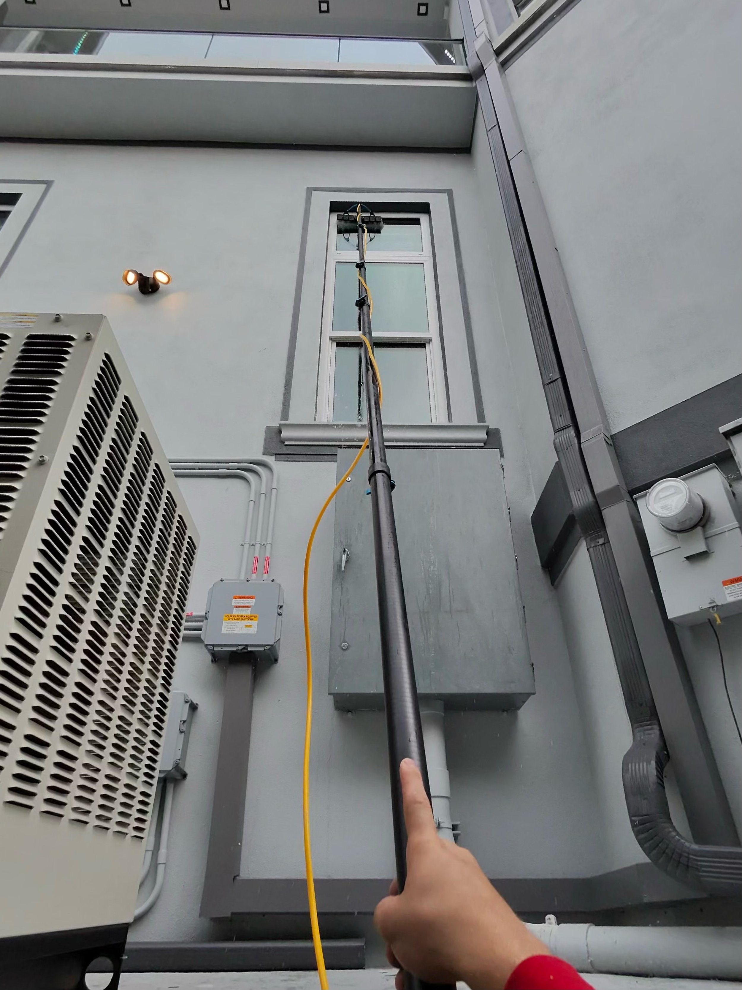Hand holding a pole reaching up to a building window, with equipment and pipes on the building exterior.