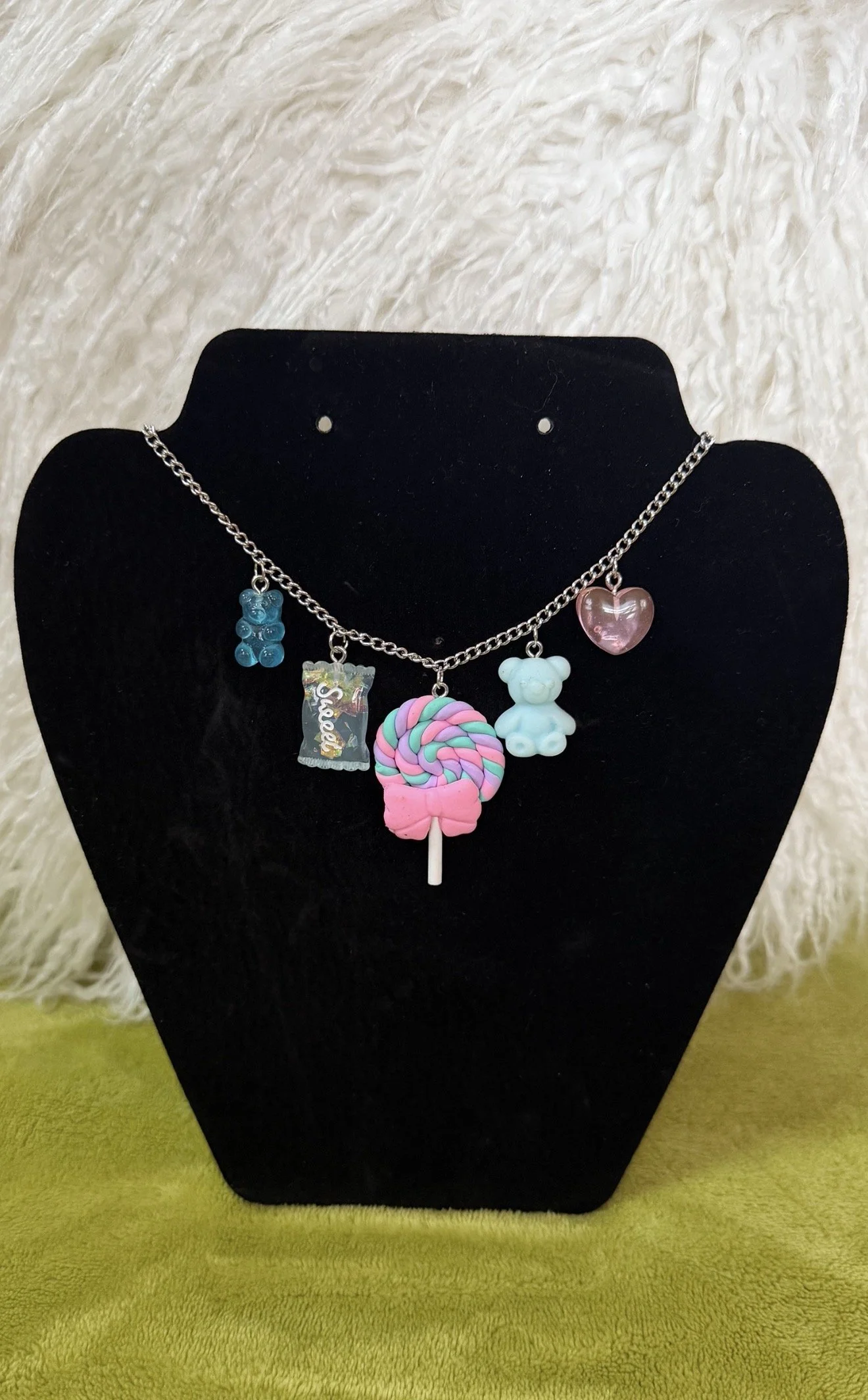 Necklace with multiple charms including a blue bear, a bag of sweets, a lollipop, a teddy bear, and a pink heart, displayed on a black stand.