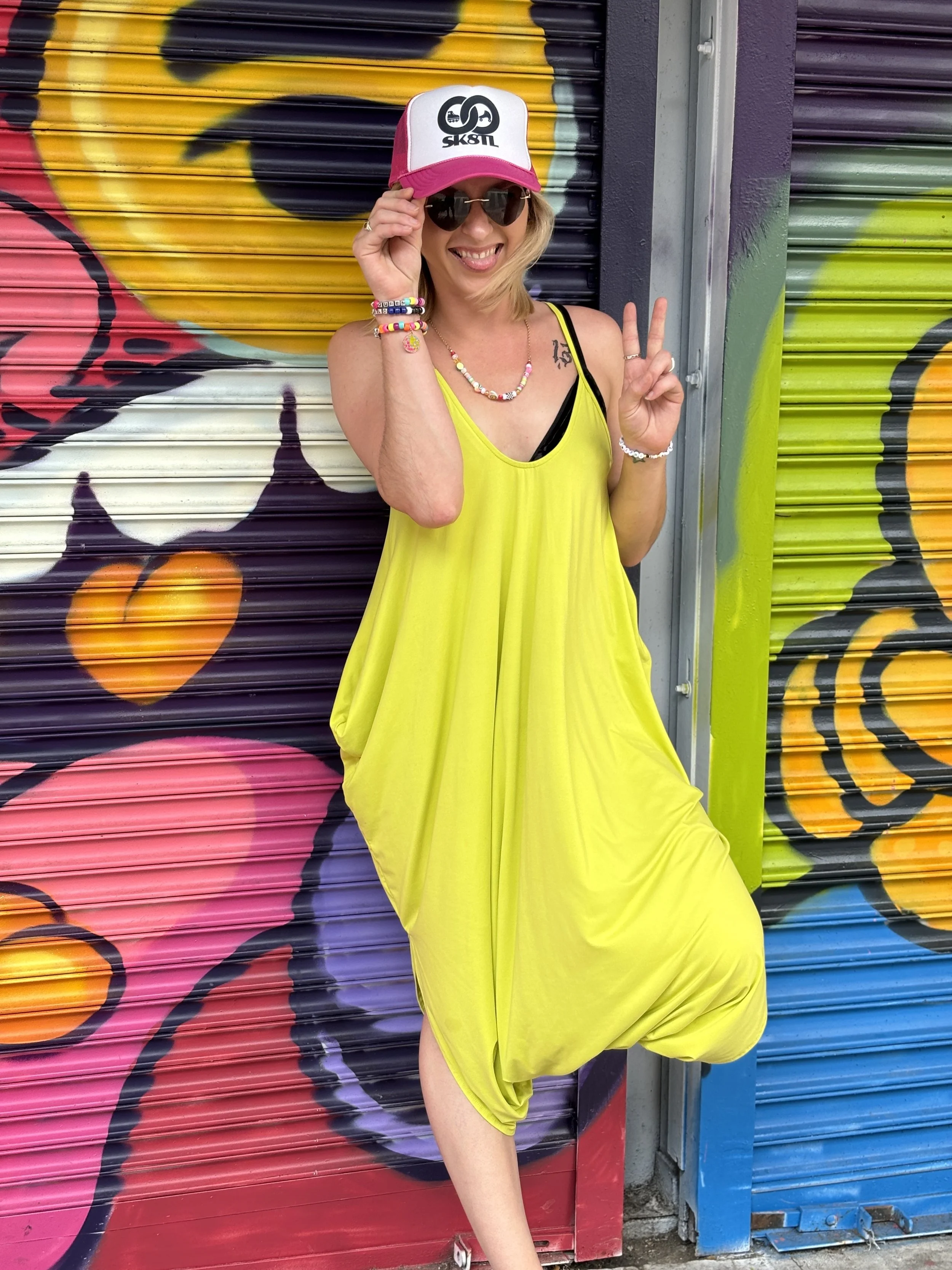 Woman in a bright yellow dress wearing sunglasses, a pink and white cap, and colorful jewelry, posing in front of a colorful graffiti mural.