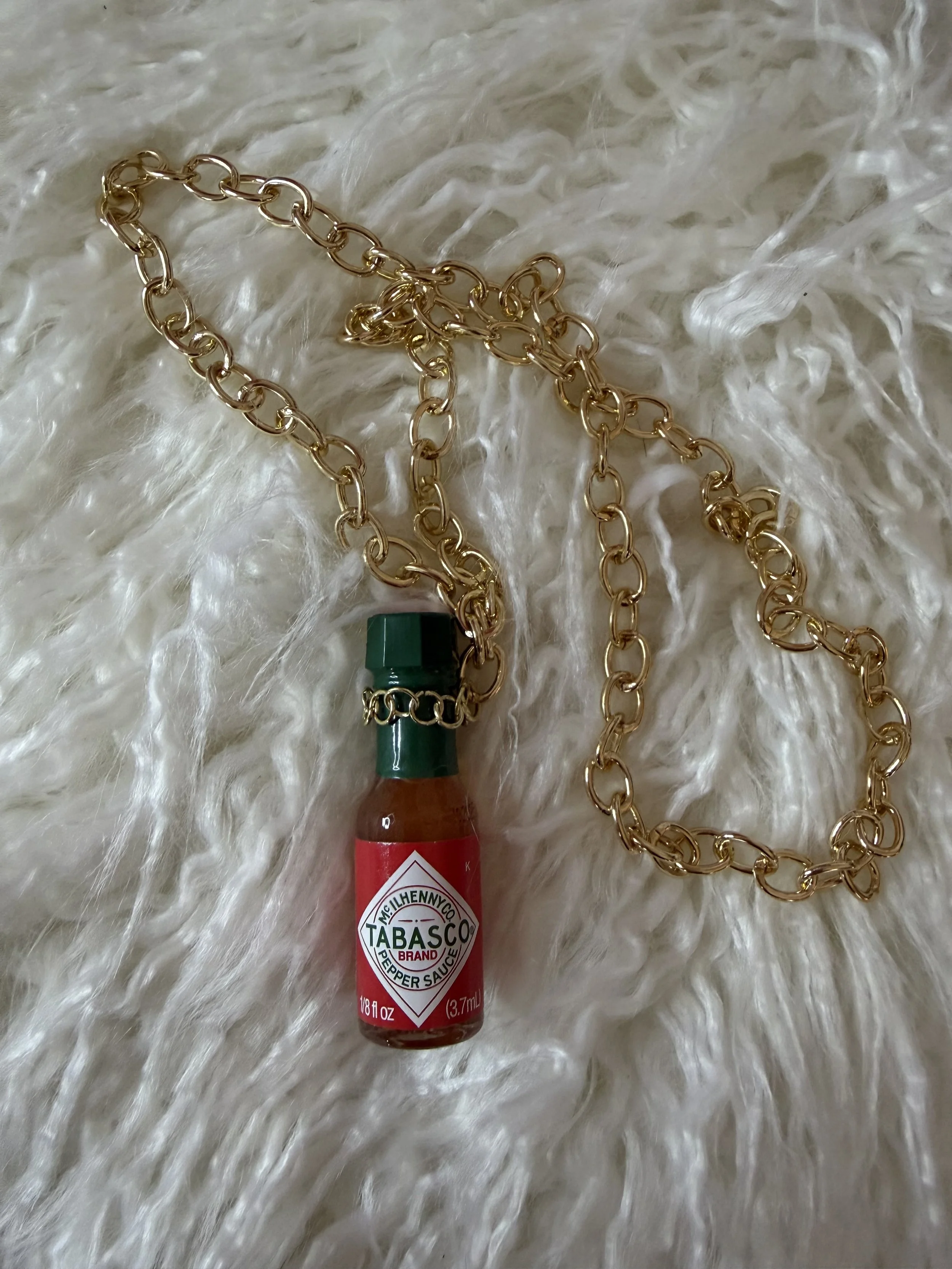 A gold chain necklace with a miniature bottle of Tabasco hot sauce attached to it, lying on a fluffy white surface.