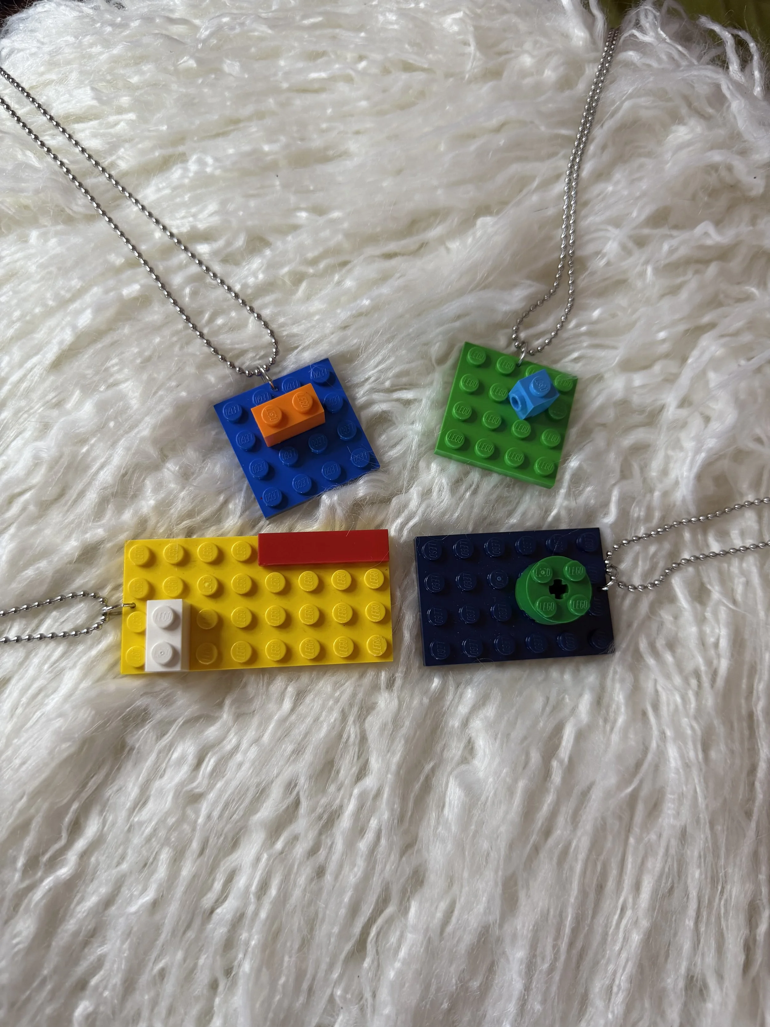 Four necklaces with pendant designs made from LEGO bricks, placed on a white furry surface.
