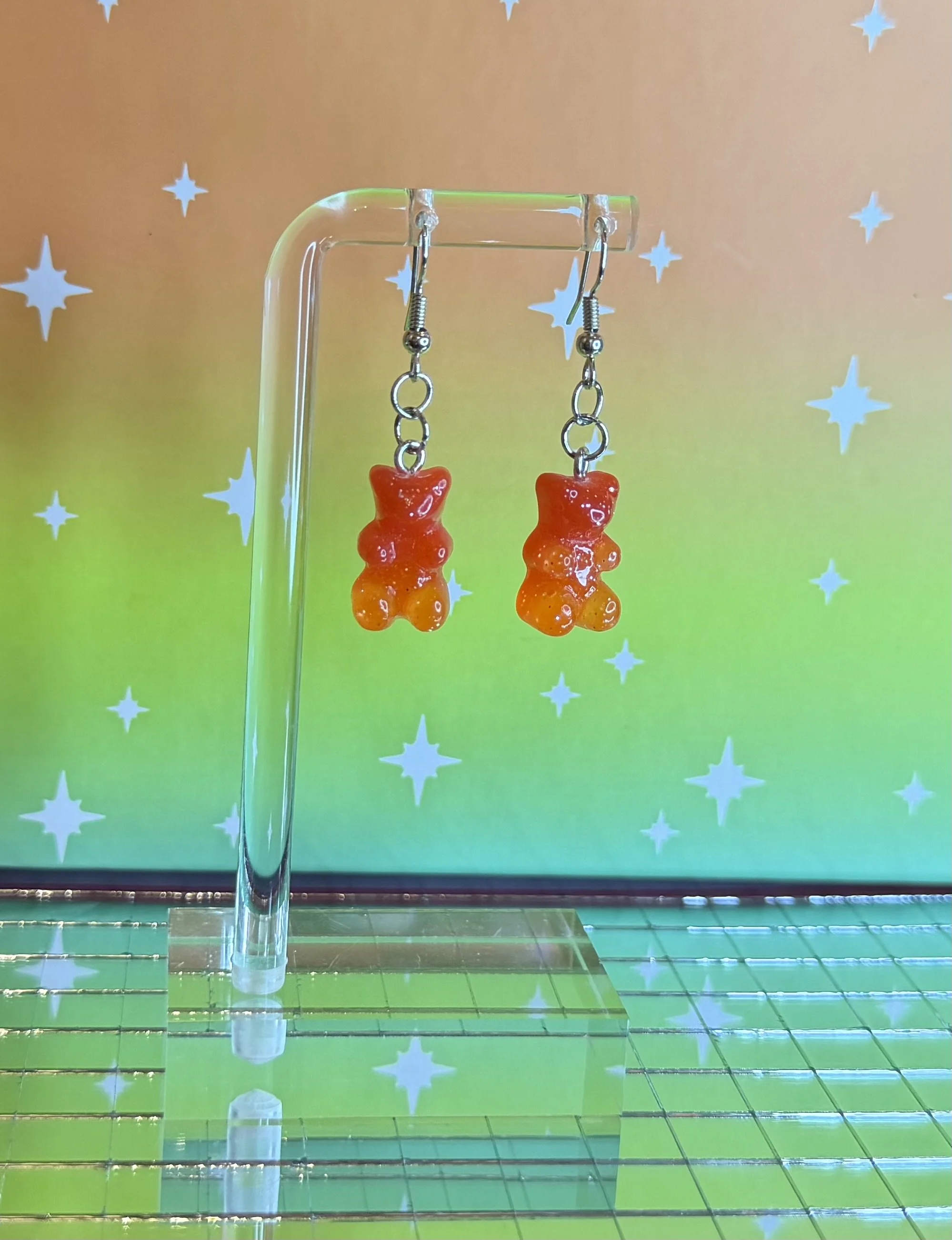Colorful gummy bear earrings hanging from a clear display stand with a starry background.