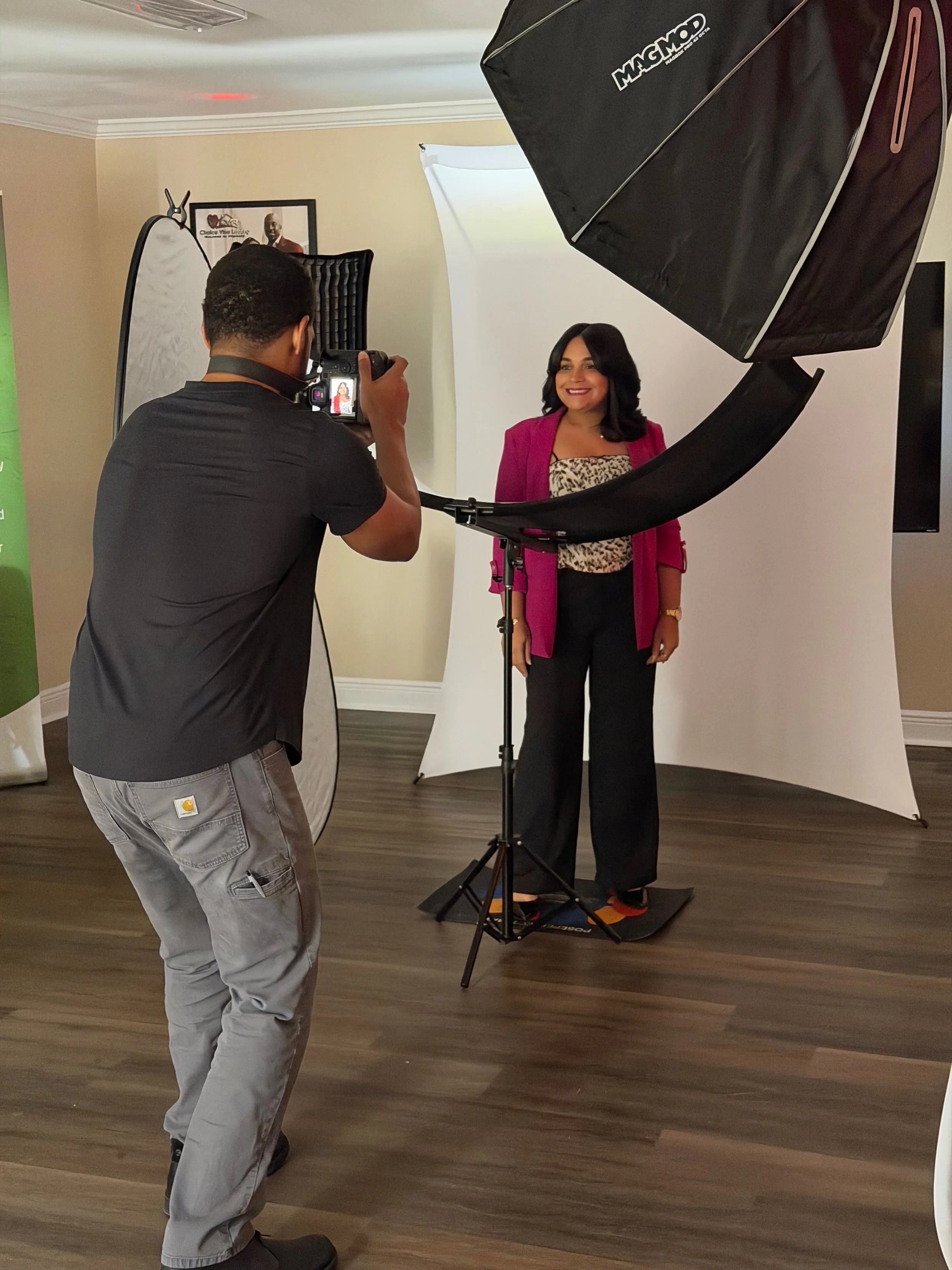 Photographer taking a portrait of a woman standing in front of a white backdrop, with professional photography equipment including softbox lights and reflectors in a studio setting.