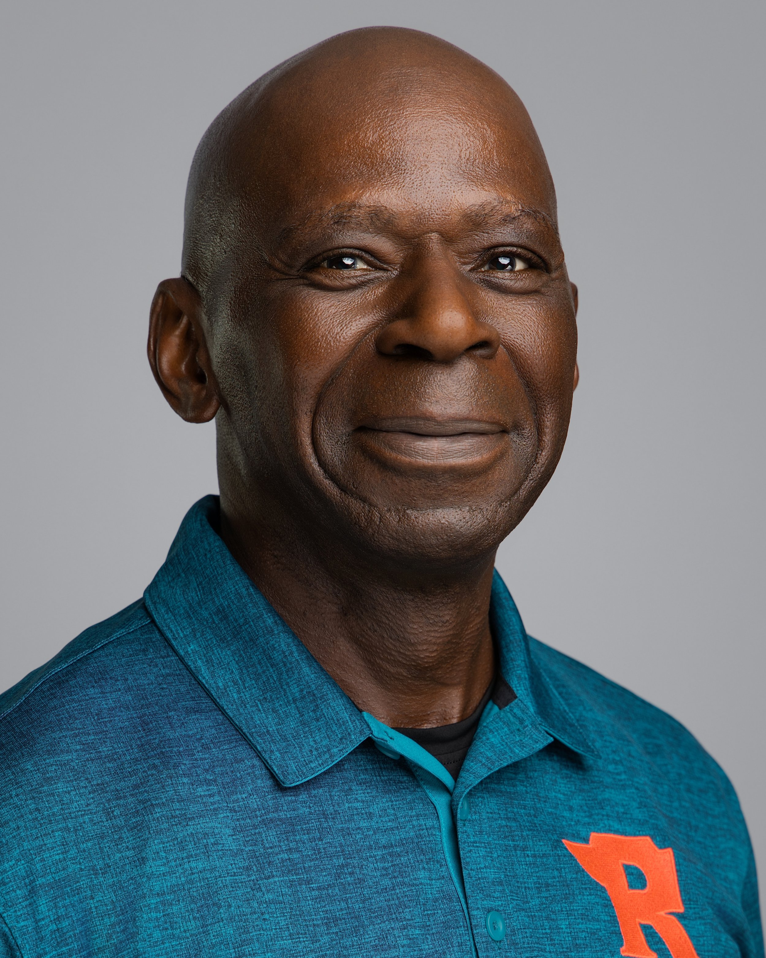 A middle-aged man with dark skin and a serious expression, wearing a blue collared shirt with an orange letter R on it, against a gray background.