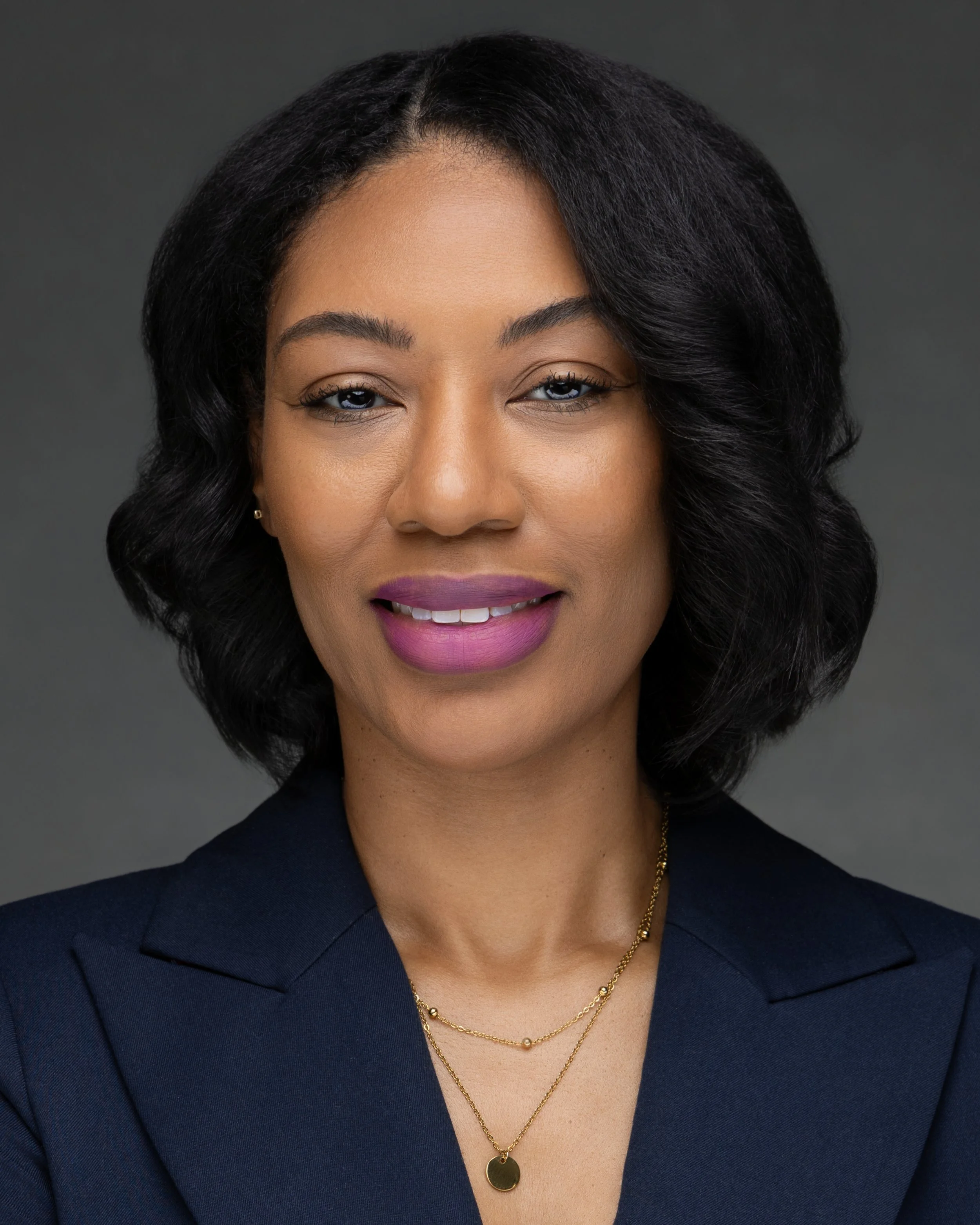 Headshot of a woman with dark,wavy hair, wearing a dark blazer, gold necklaces, light makeup, and purple lipstick, against a gray background.