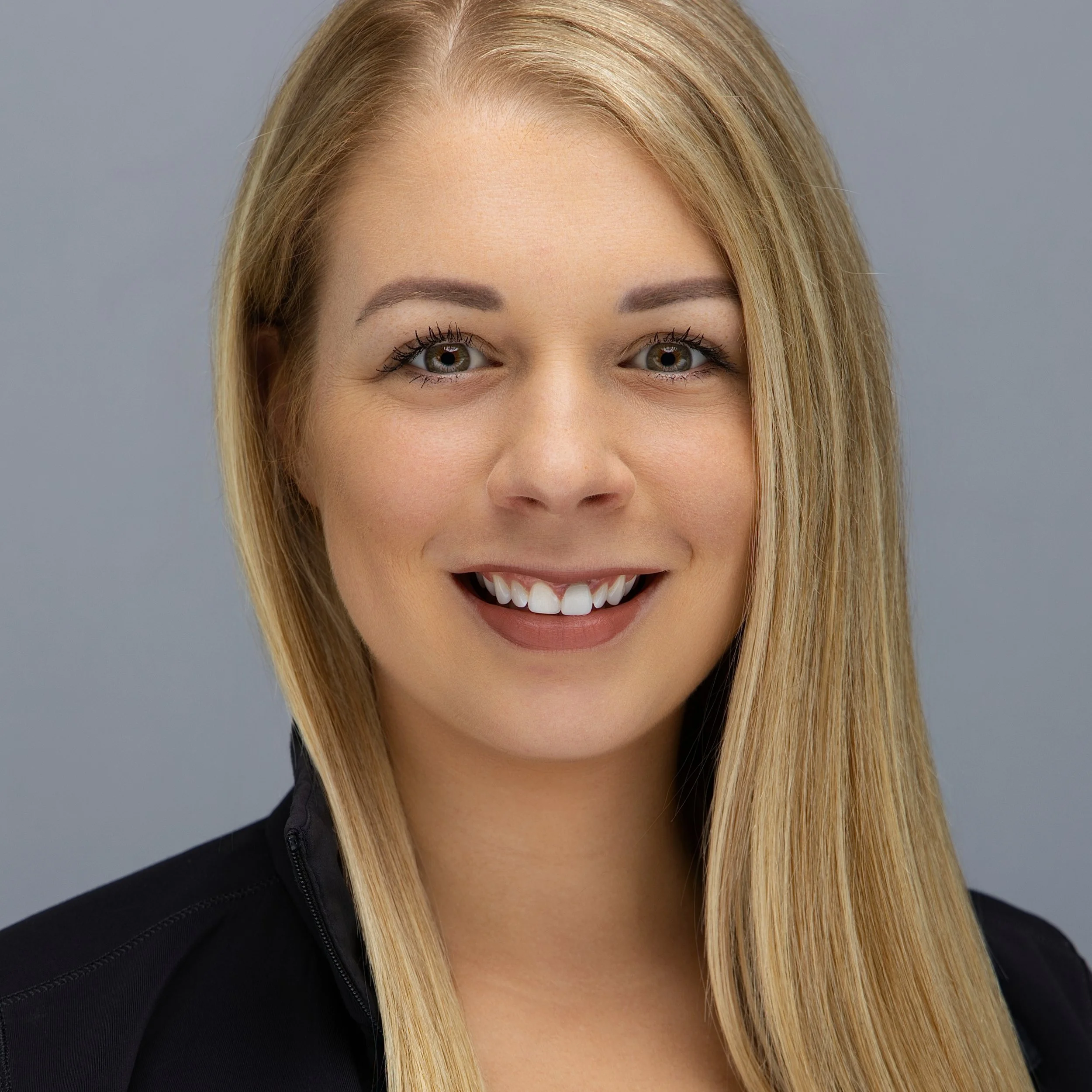 Close-up portrait of a young woman with blonde hair, smiling, wearing a black top, against a gray background.