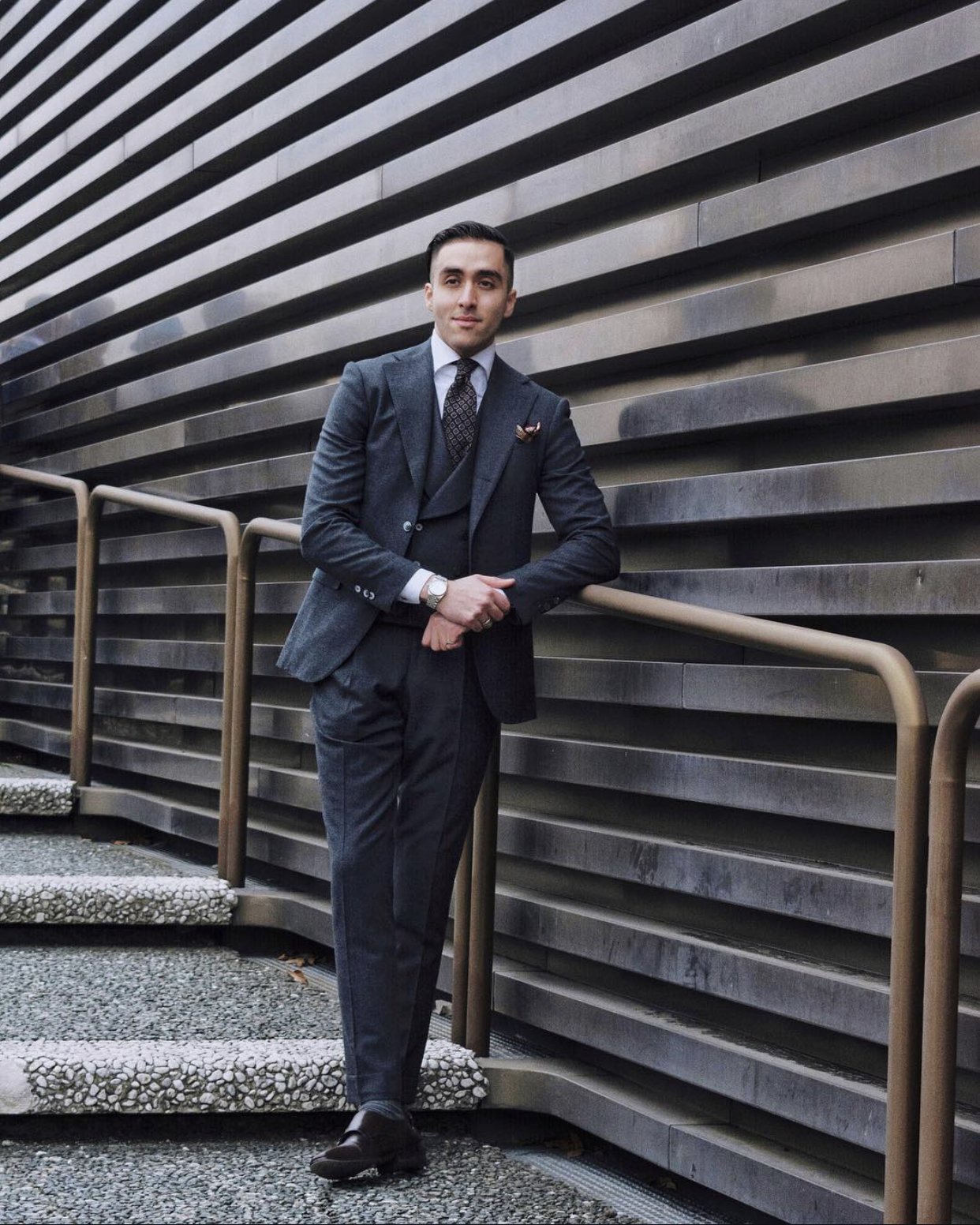 A man in a dark gray suit with a white dress shirt, patterned black tie, and a watch, standing outdoors next to a railing, in front of a modern building with horizontal metal louvers.