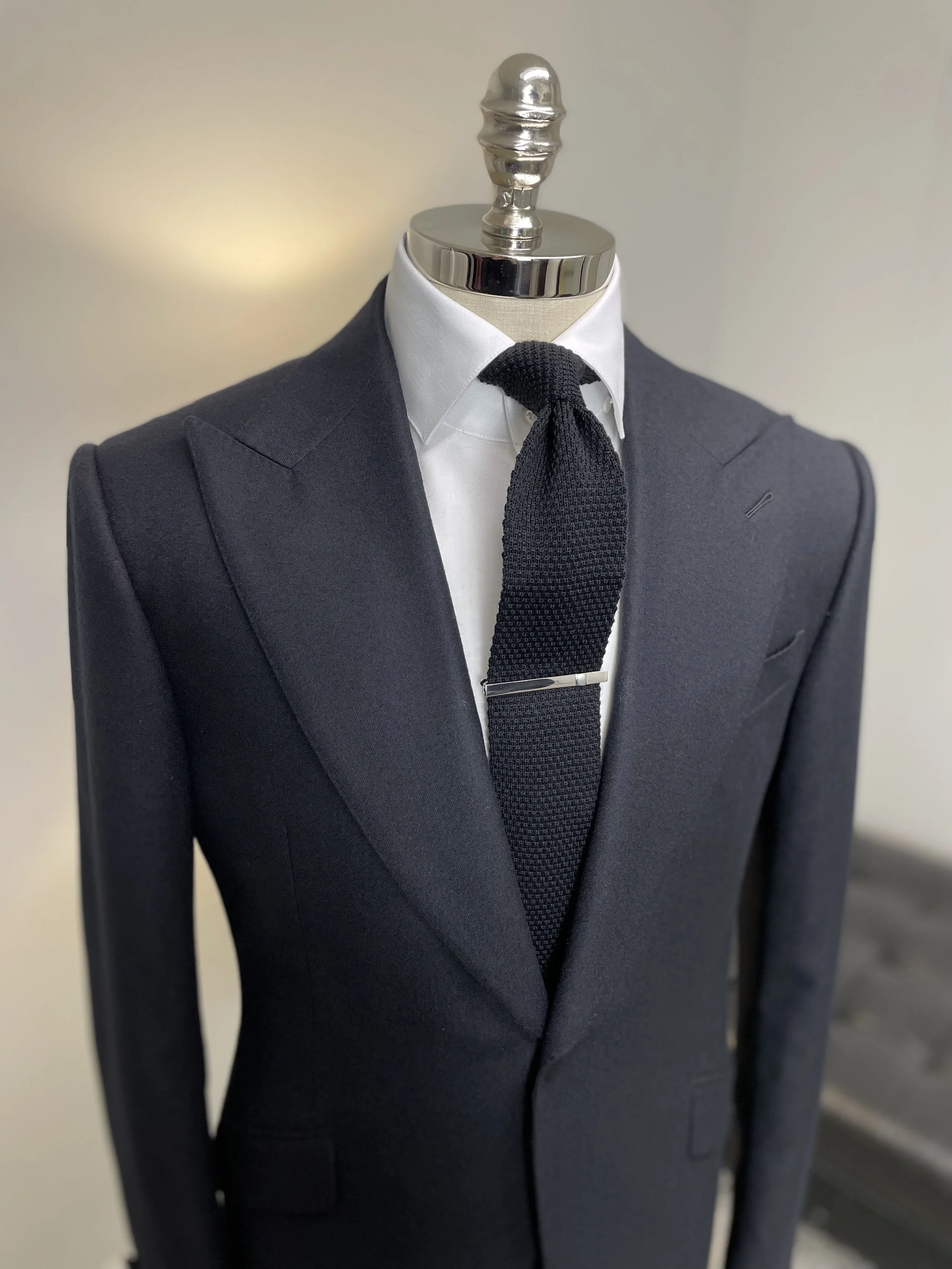 Close-up of a mannequin dressed in a dark suit jacket, white dress shirt, and black textured tie with a tie clip.