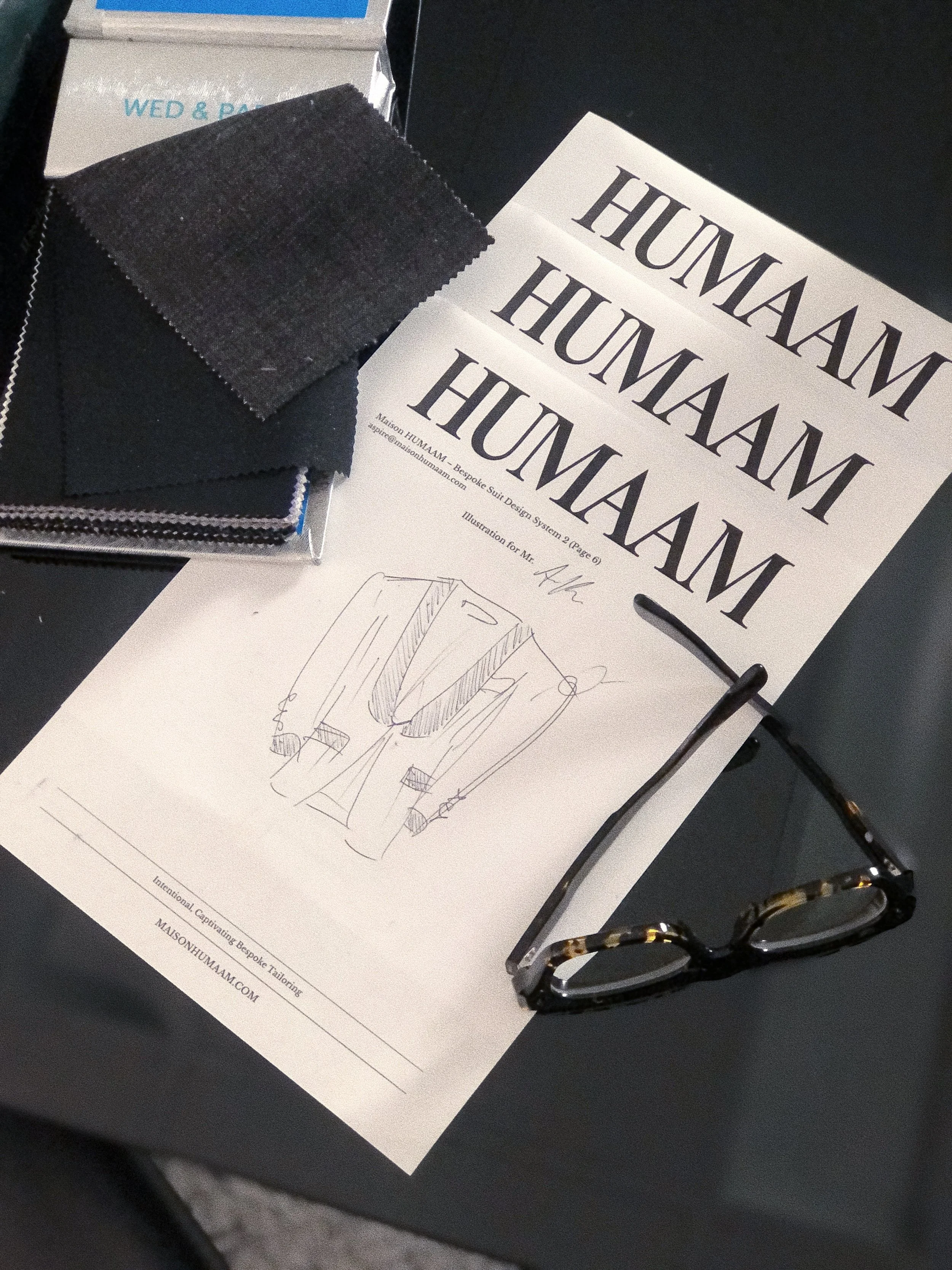 Fabric swatches, designer sketches, glasses, and printed materials on a black surface related to high-end fashion design.