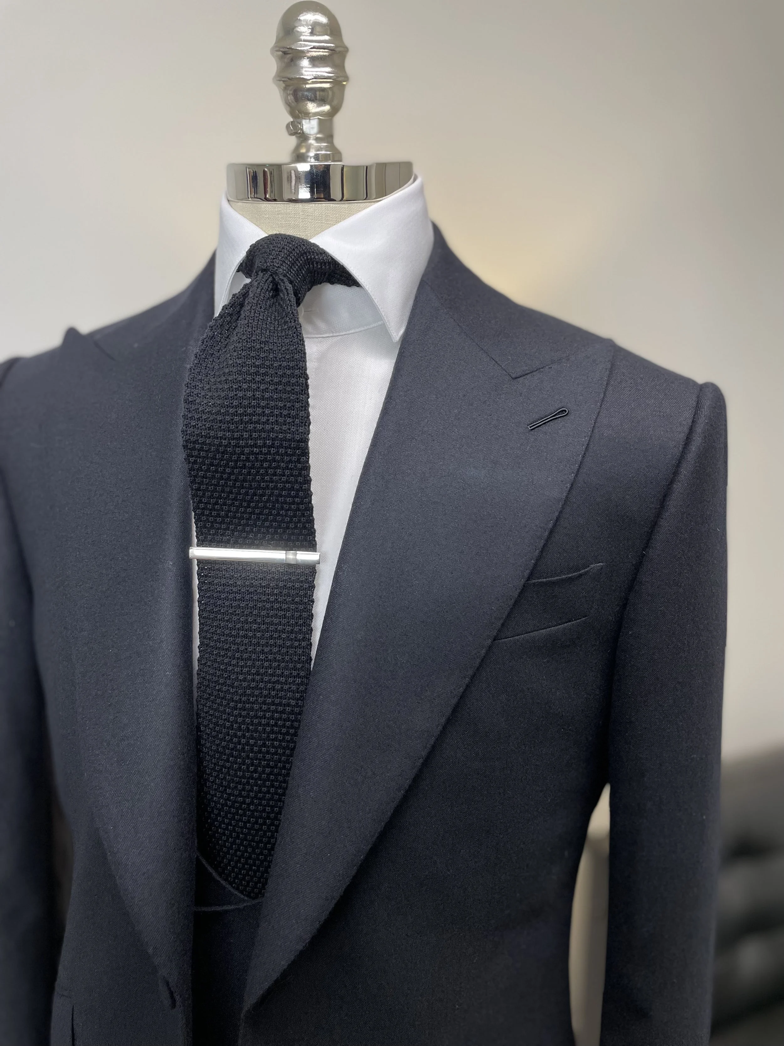Formal men's suit display with a dark blazer, white shirt, black textured tie, and tie clip on a mannequin