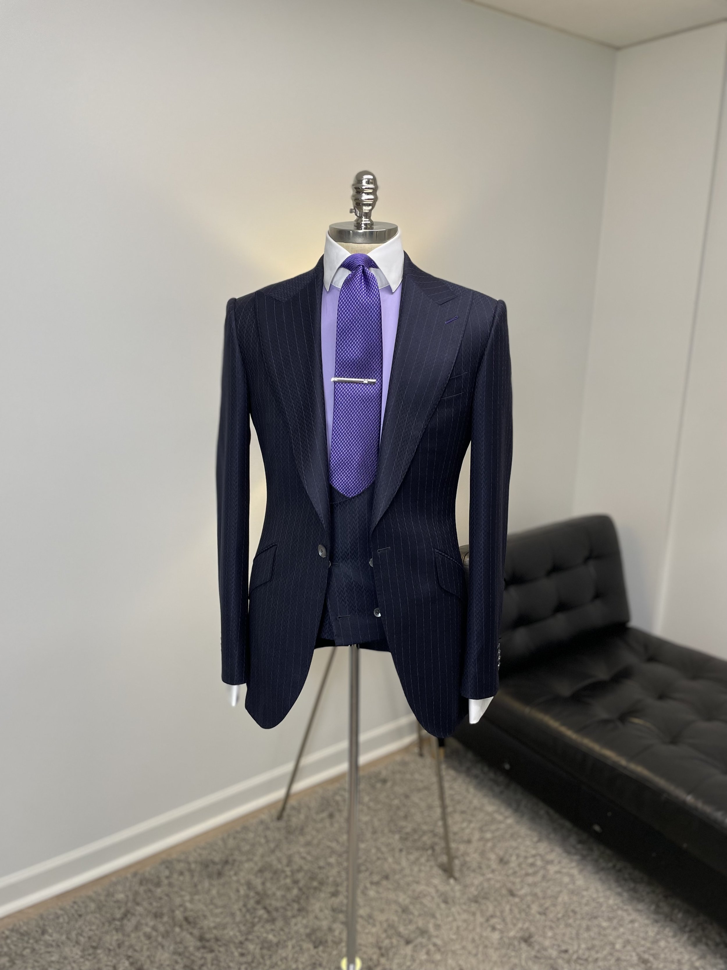 A mannequin dressed in a dark pinstripe suit, white shirt, purple tie, and waistcoat, with a silver tie clip, displayed on a metal stand against a plain wall, with a black leather sofa in the background.