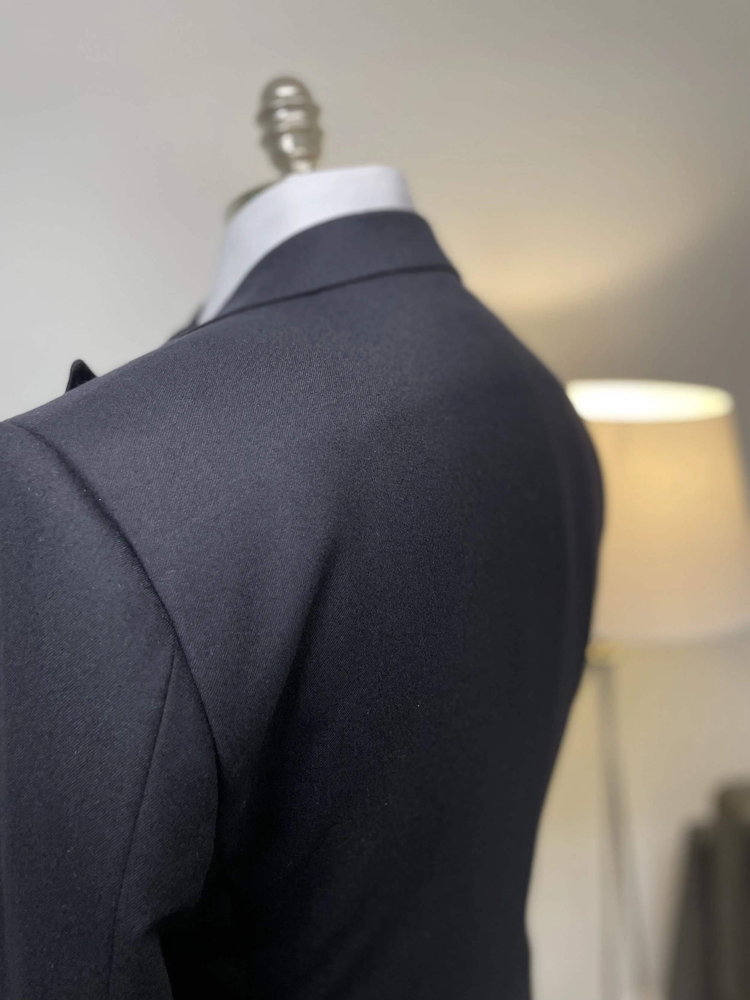 Close-up of a dark suit jacket on a mannequin with a white collar, in a room with a lamp and a beige wall in the background.