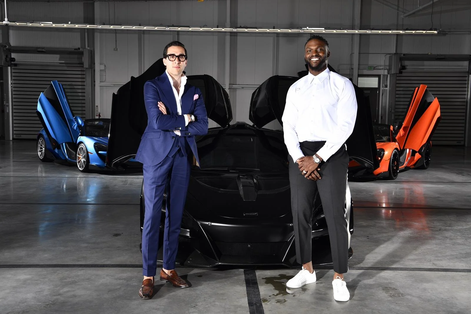 Two men (Humaam and Cliff Avril) standing in front of a black sports car, with two additional sports cars with open doors in the background, inside a garage or showroom.