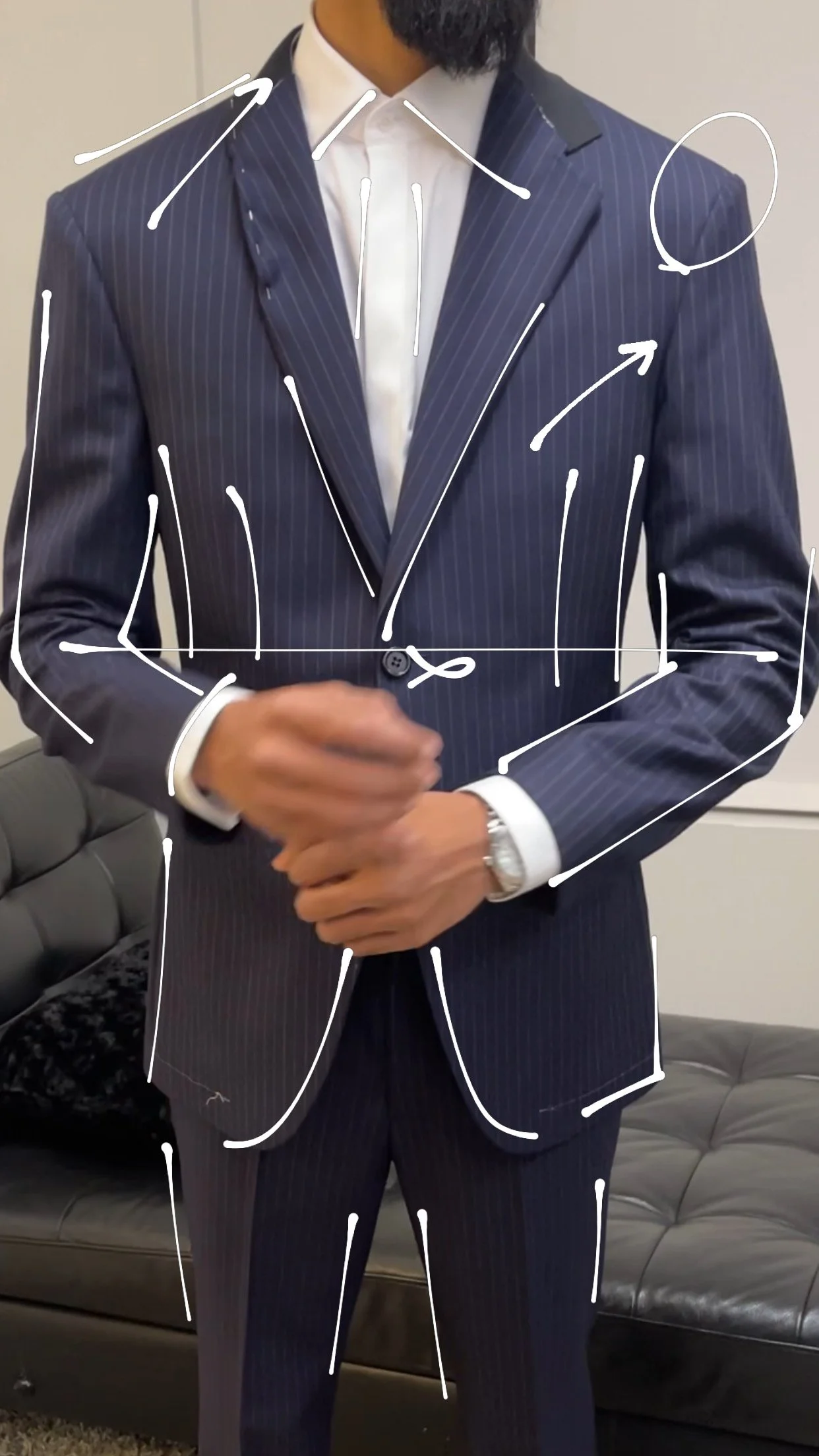 Man wearing a navy blue pinstripe suit with a white shirt, adjusting his suit jacket, sitting on a gray leather couch in an indoor setting.