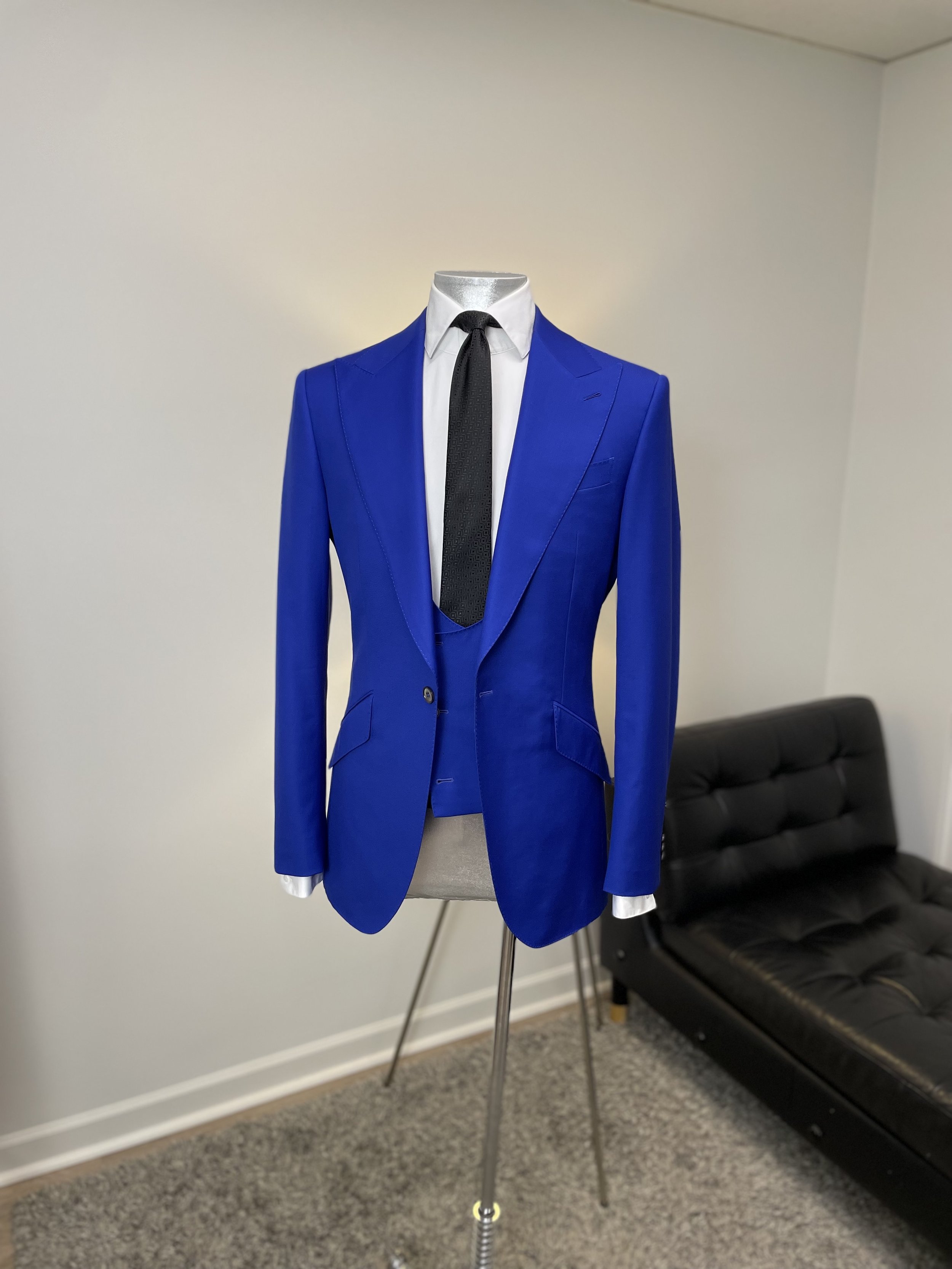 A mannequin wearing a bright blue suit with a white shirt and black tie, displayed on a stand in a room with beige carpeting and a white wall, next to a black leather sofa.