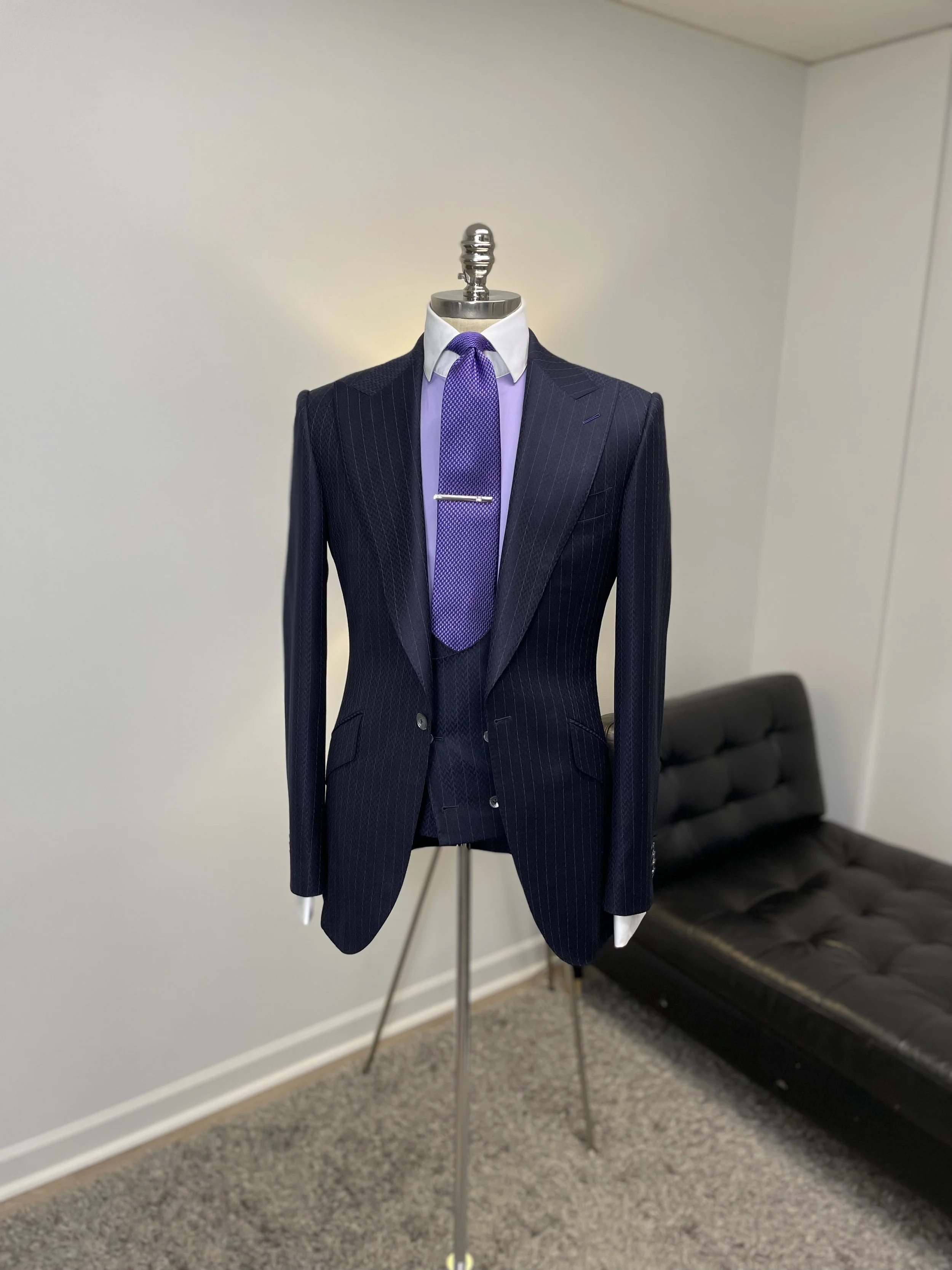 A mannequin dressed in a navy pinstripe suit with a white shirt, purple tie, and tie clip, displayed in an indoor room with white walls, next to a black tufted sofa.