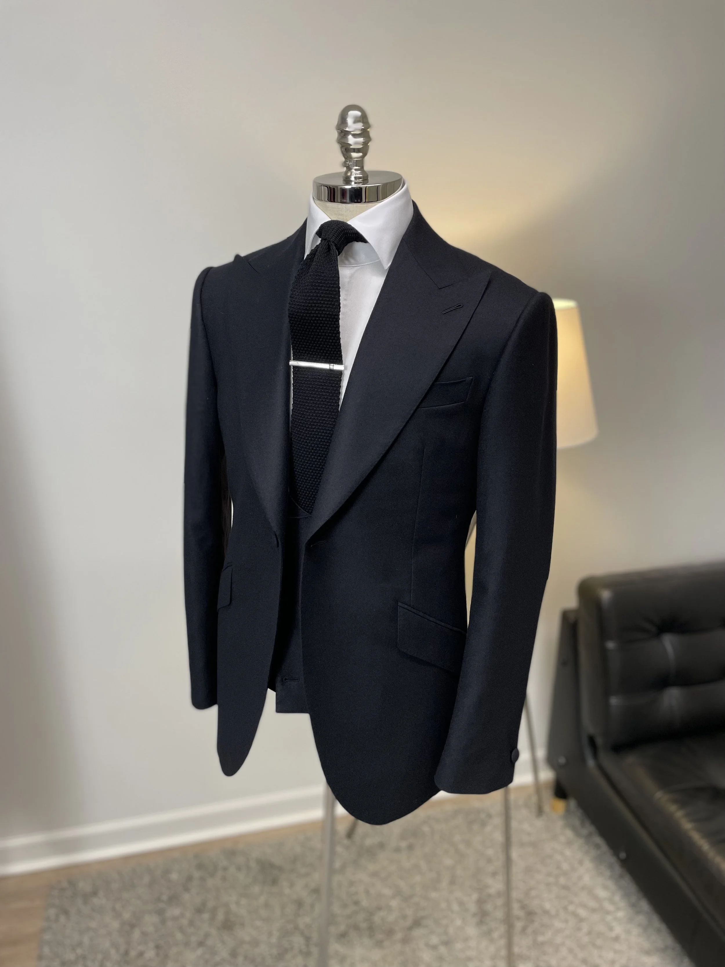 A black suit with a white shirt and a black textured tie on a mannequin, standing in a room with a black leather chair and a lamp.